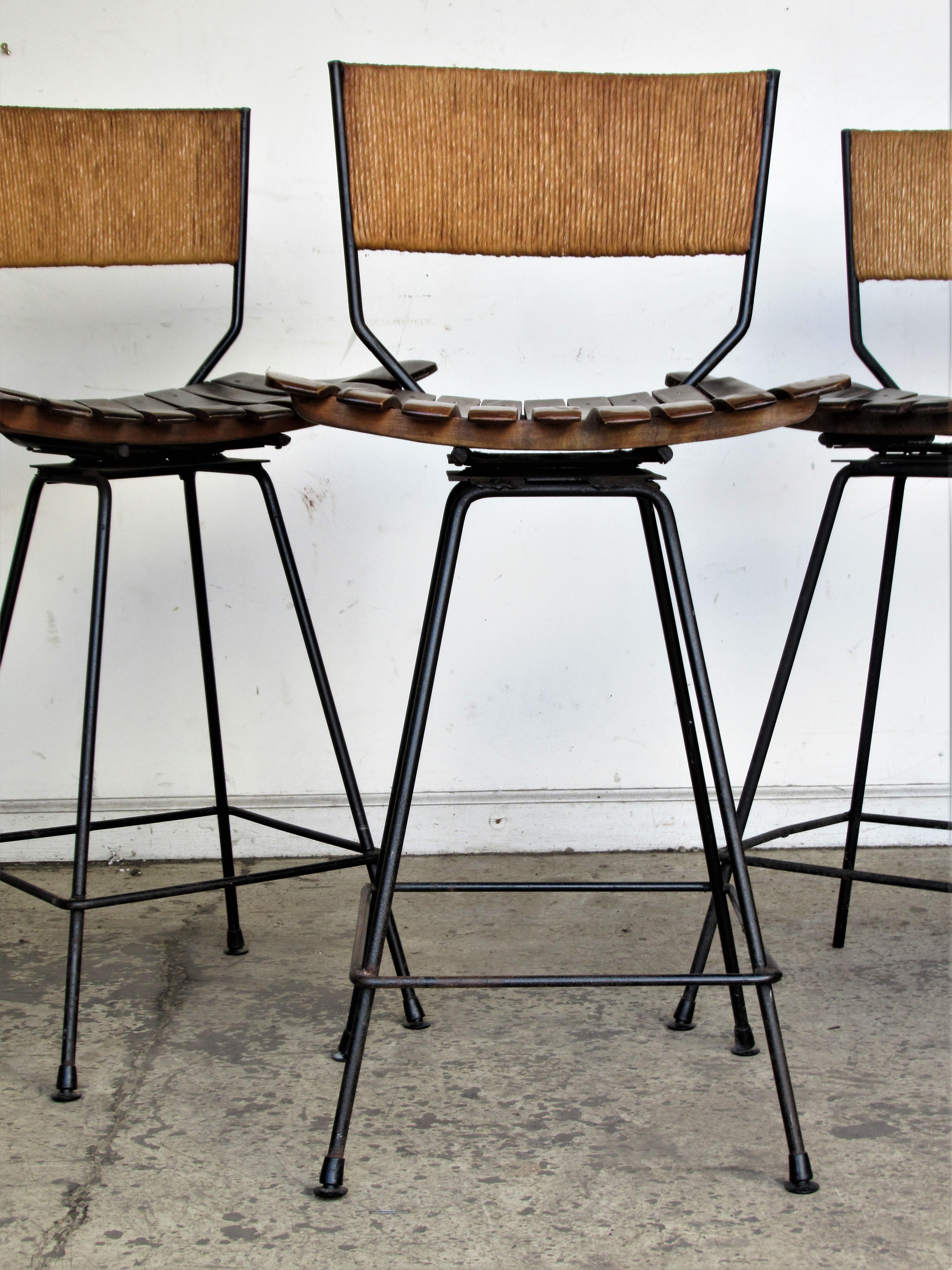 Set of three matching mid-20th century modern swivel seat stools with black iron frames, woven rush backs and contoured wood plank seats in great original vintage condition by Arthur Umanoff for Raymor, circa 1960. Look at all pictures and read