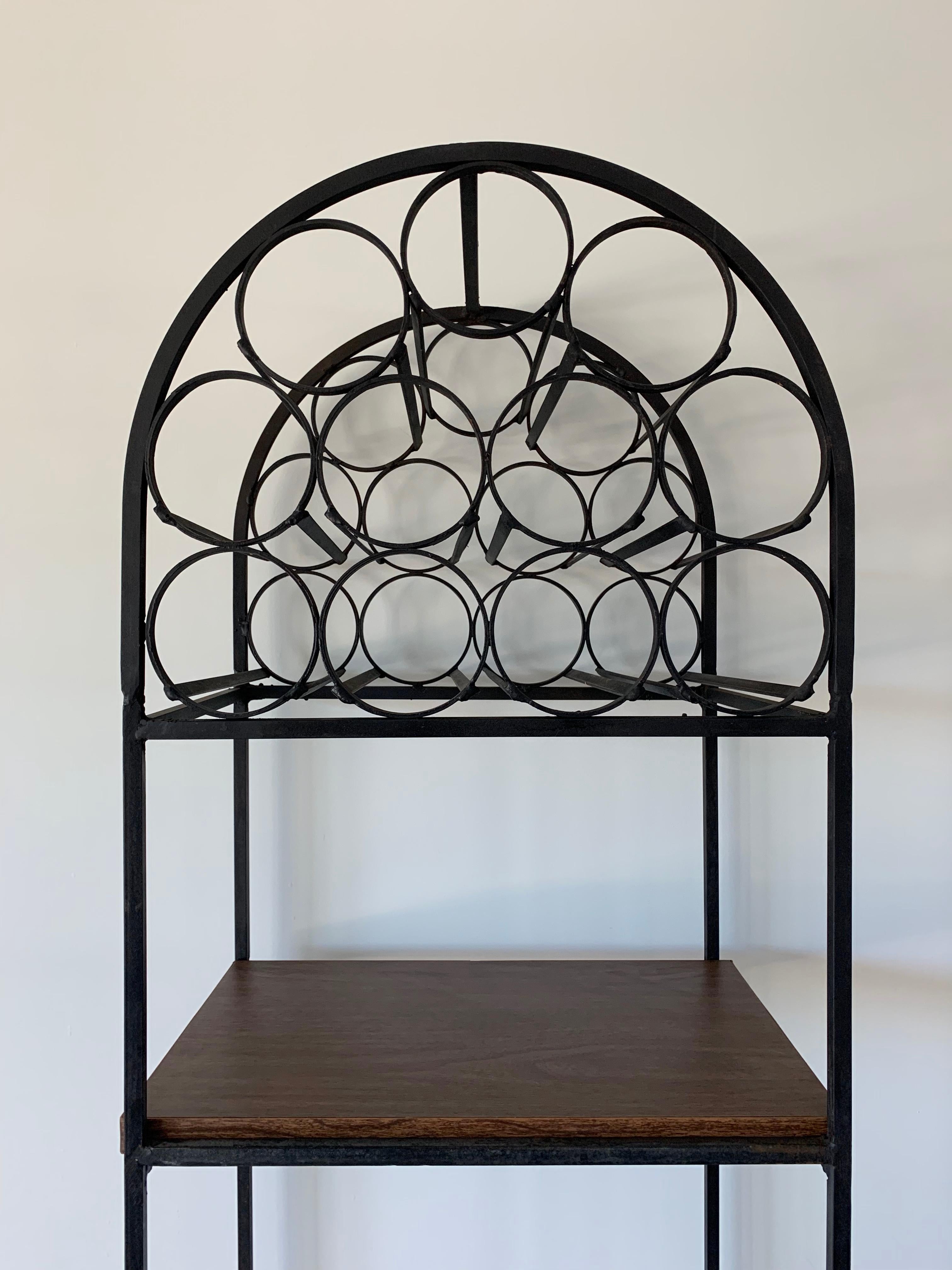A black wrought iron wine rack by Arthur Umanoff.
It can hold up to 39 bottles and includes a woven basket type shelf and a laminated shelf to hold glasses, serve drinks or even showcase you favorite bottles and bar utensils.
Manufactured for the