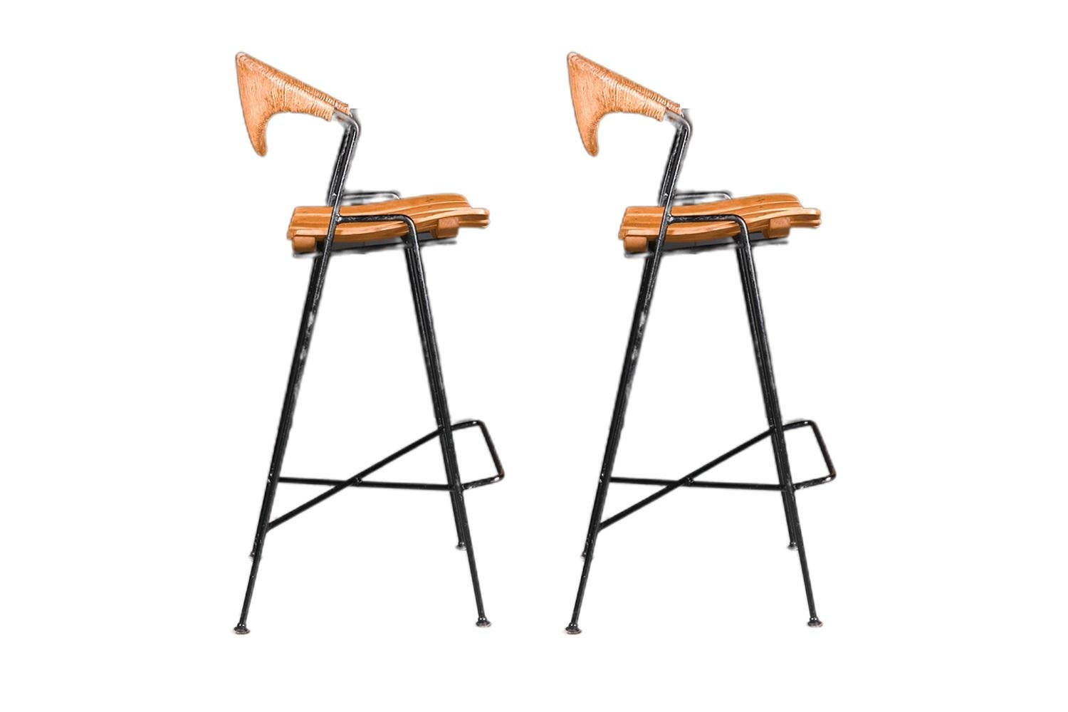 A Pair of Mid Century paper cord and slat seat bar height stools, with very thin iron bases, designed by Arthur Umanoff for Shaver Howard each remain in original condition throughout. Features stylish, sculptural, curved paper cord backrests in a