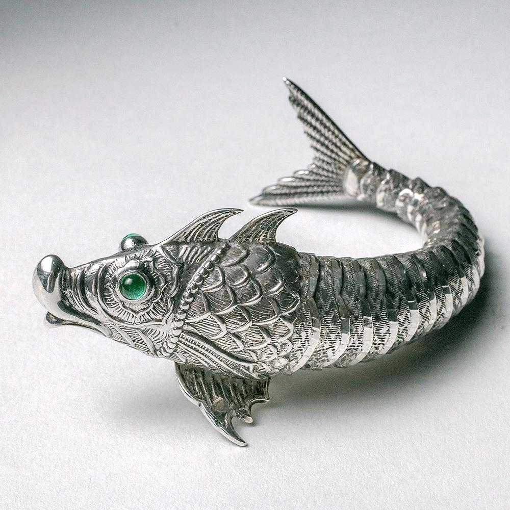 A smaller vintage Spanish sterling silver fish resting on two lower pectoral fins with fully articulated body, two green glass eyes, and tail, all masterfully engraved. The body is beautifully crafted in articulated sections that give the fish