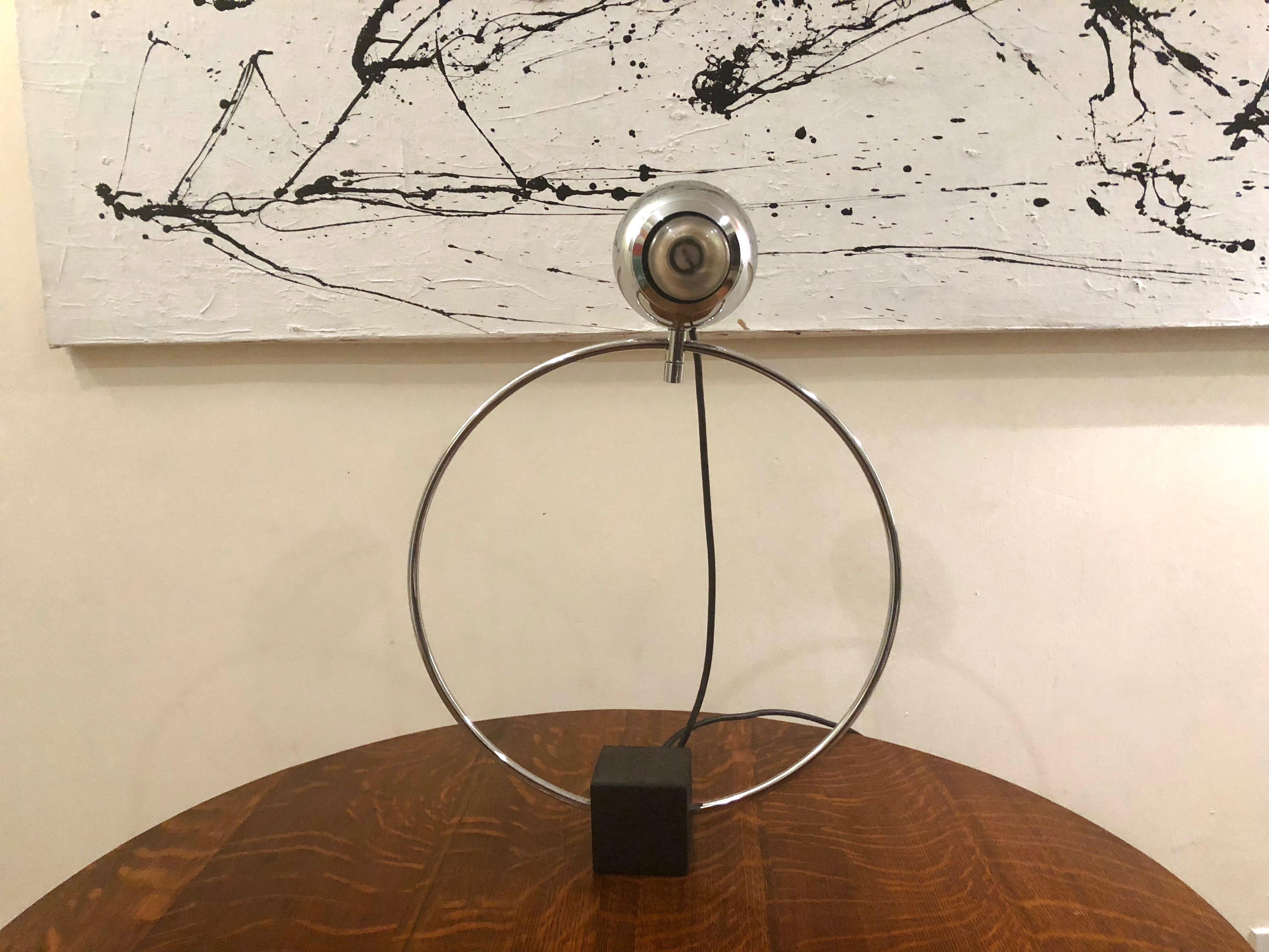 La lampe de table / de bureau est composée d'une base cubique en acier noirci qui supporte un cadre circulaire en métal chromé poli et un diffuseur de forme atomique également en métal chromé poli. Le diffuseur est une tête sphérique articulée à 360