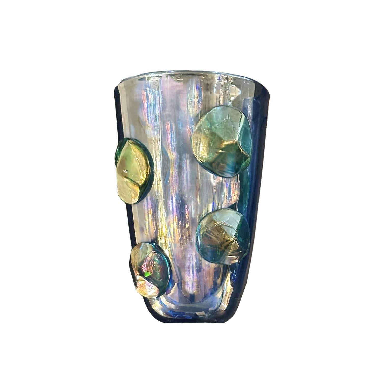 Questo vaso di grande impatto visivo utilizza colori vivaci e iridescenti e bottoni di vetro applicati per aggiungere dimensione e profondità alla superficie del vetro. I vortici di viola, verde e blu che si muovono al variare della luce renderanno