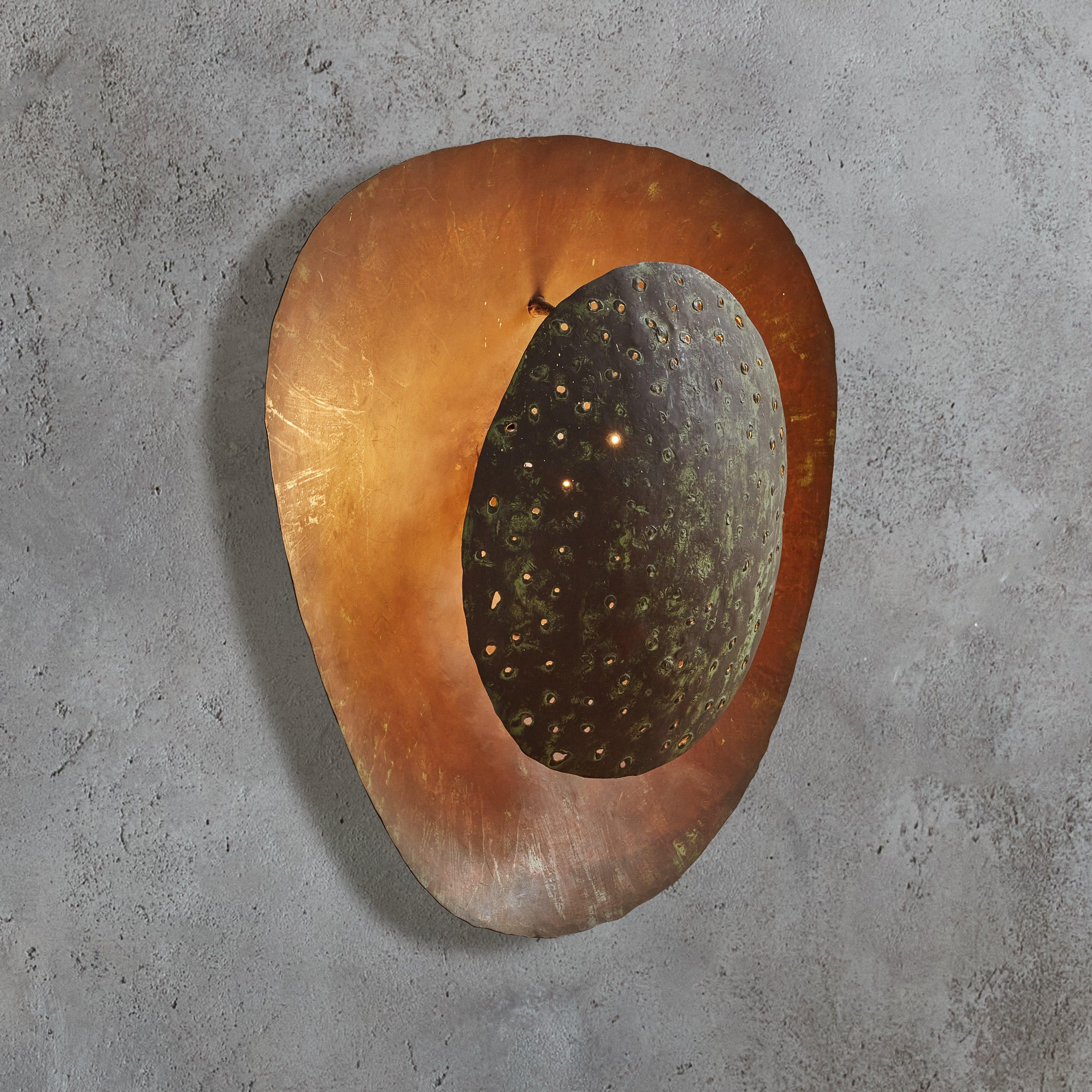 A rare French artisan made metal sconce from the 1950s. The sculptural, organic modern silhouette is composed of two layered patinated metal discs with delicate holes and rich glazing in deep, earthy hues. When lit, shades of olive green, plum, and