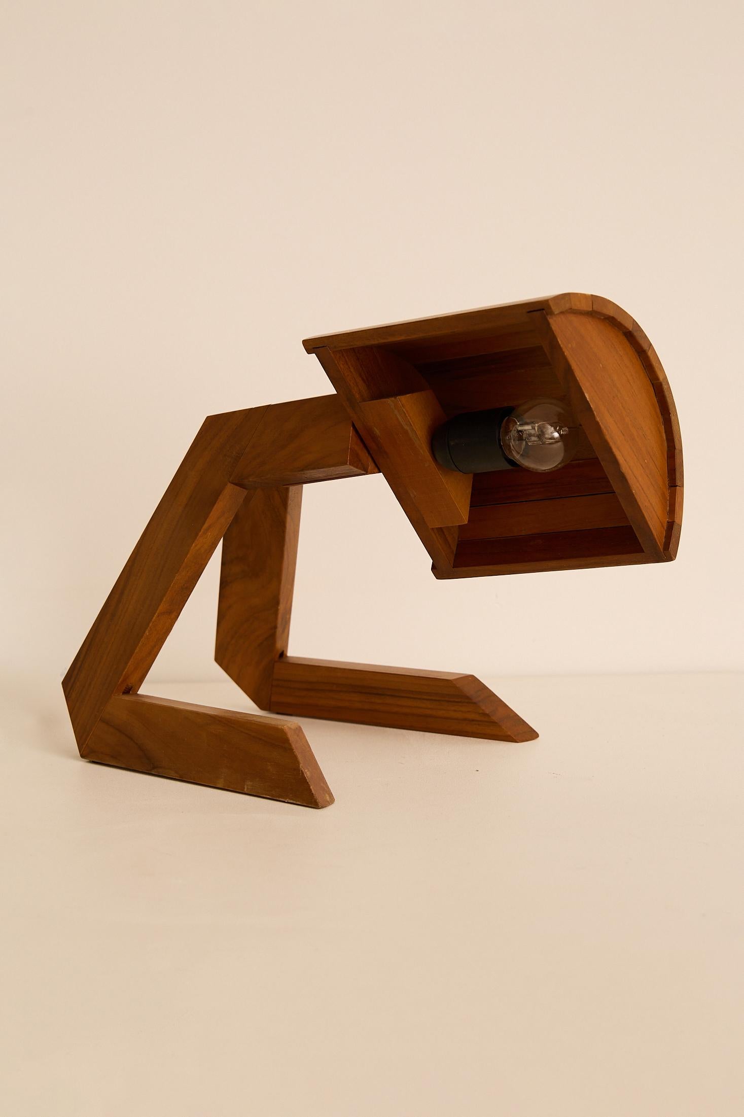 Lámpara de mesa artesanal de madera con una base escultural y angular y una pantalla de medio cilindro que dirige un resplandor cálido y concentrado. Hecho a mano con madera maciza, combina líneas arquitectónicas con una luz suave y atmosférica.