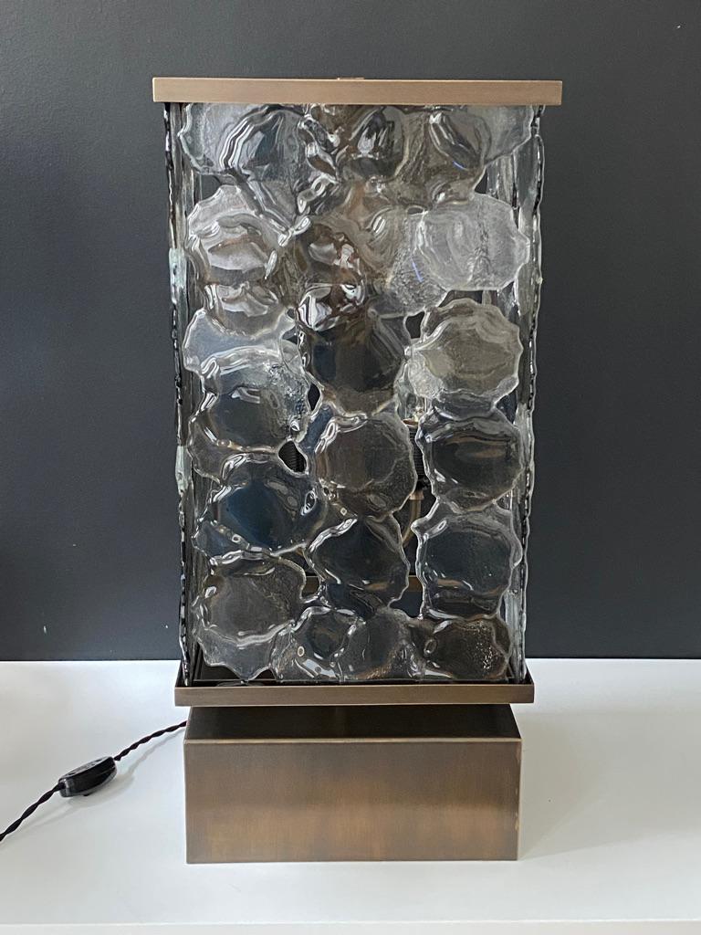 Truly a unique piece with fused hand worked glass panels. The glass panels are a mix of bronze, gray and clear glass that would complement any interior. 
The metal base, woven electric cord and the fabric shade, all add to its timeless character.