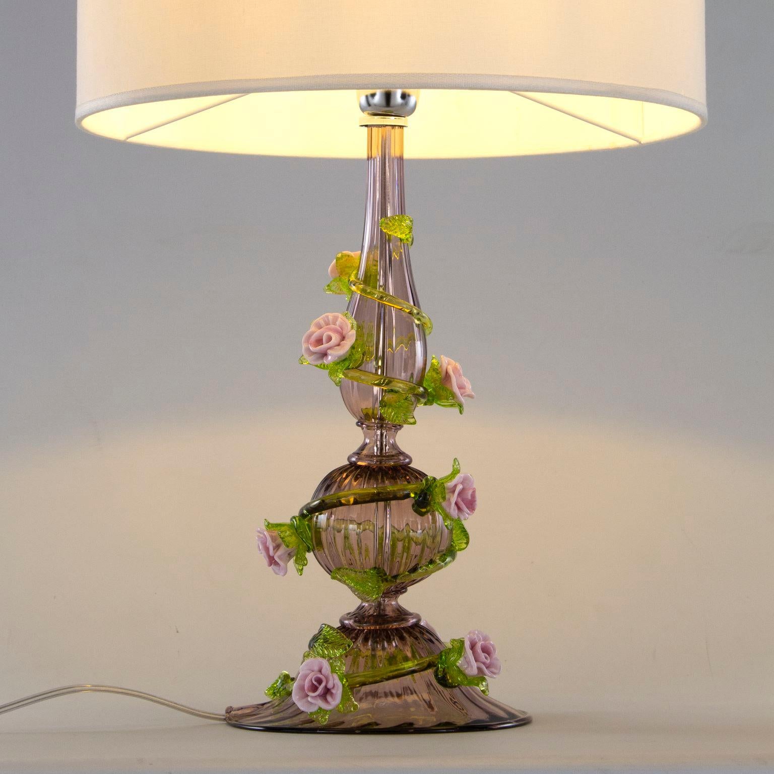 Rosae Rosarum Garden is a table lamp with 1 light, in amethyst colour and details in green and in pink vitreous paste. A white fabric lampshade with a rectangular shape covers the light bulb. It is a romantic and with natural elements table lamp.