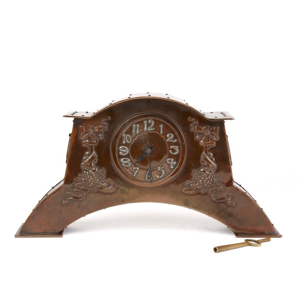 An exceptional Arts & Crafts copper mantel clock standing on two rectangular shaped feet with an arched body with an embossed dial and silvered Arabic numerals flanked with ribbons and garlands of fruit the arched case with riveted plates and