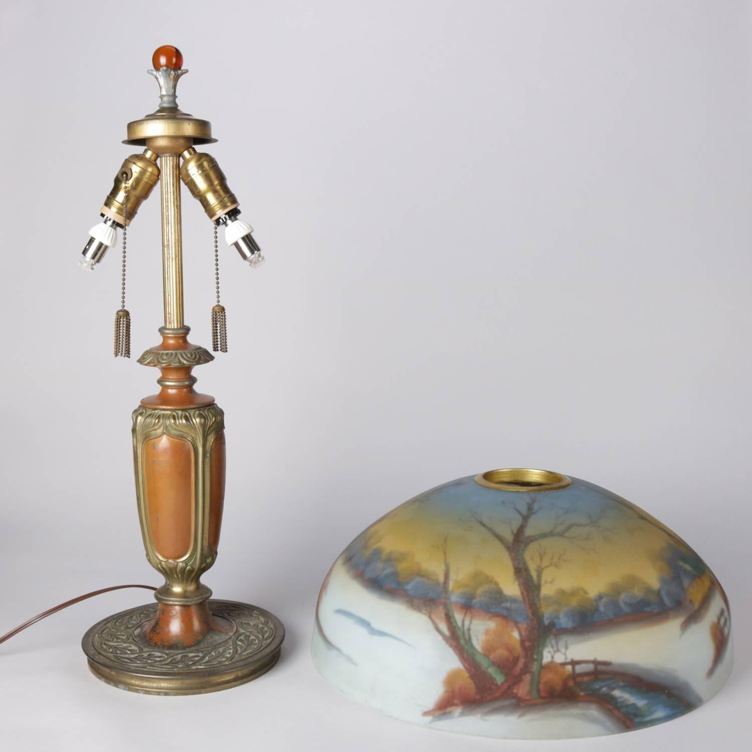 Arts & Crafts Pittsburgh School reverse painted table lamp features coppered and gilt foliate decorated cast base with dual lights and reverse painted shade depicting winter landscape stream scene with footbridge, finial with amber glass bead, newly