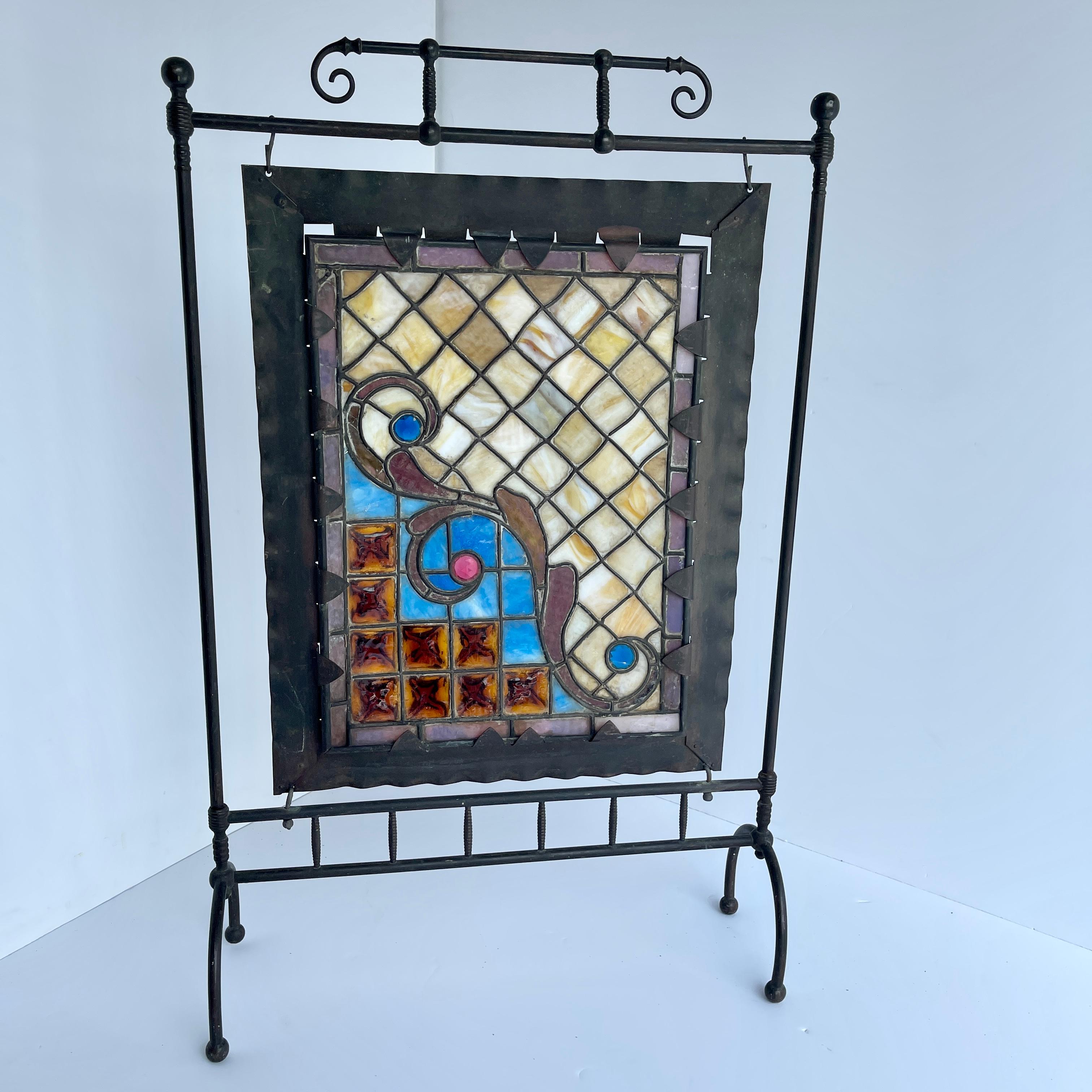 Iron fireplace screen with stained glass, Arts & Crafts era.
The unique design of this fireplace screen is enhanced by the light shining through the colorful stained glass. The center is framed in black metal with decorative curved iron braces. The