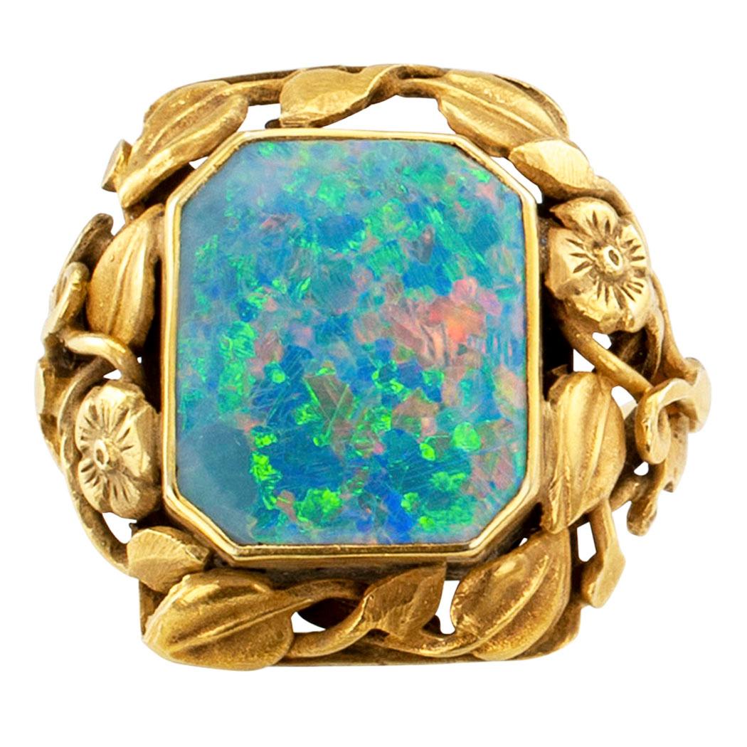 
Arts and Crafts black opal and gold ring circa 1900.  Showcasing a bezel-set, octagonal natural black opal framed by a lavish, winding garland of flowers and leaves to the shoulders and shank, mounted in 14-karat yellow gold with a matt finish