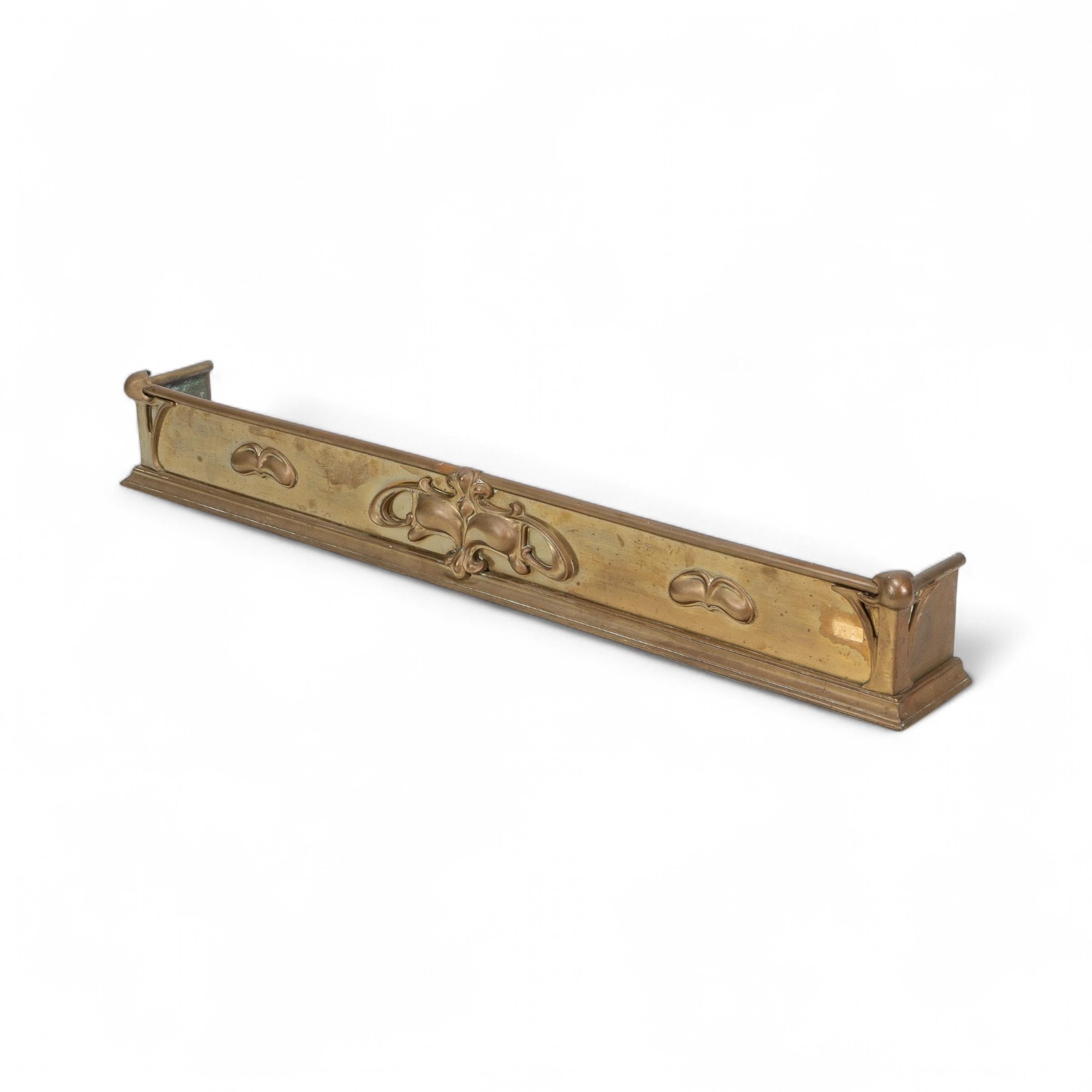 An Arts and Crafts brass fender with embossed floral decoration featuring a central stylised flower flanked by softly rounded organic forms. The front is bordered with raised edges and bracket-like corners, all cast in relief with a naturalistic