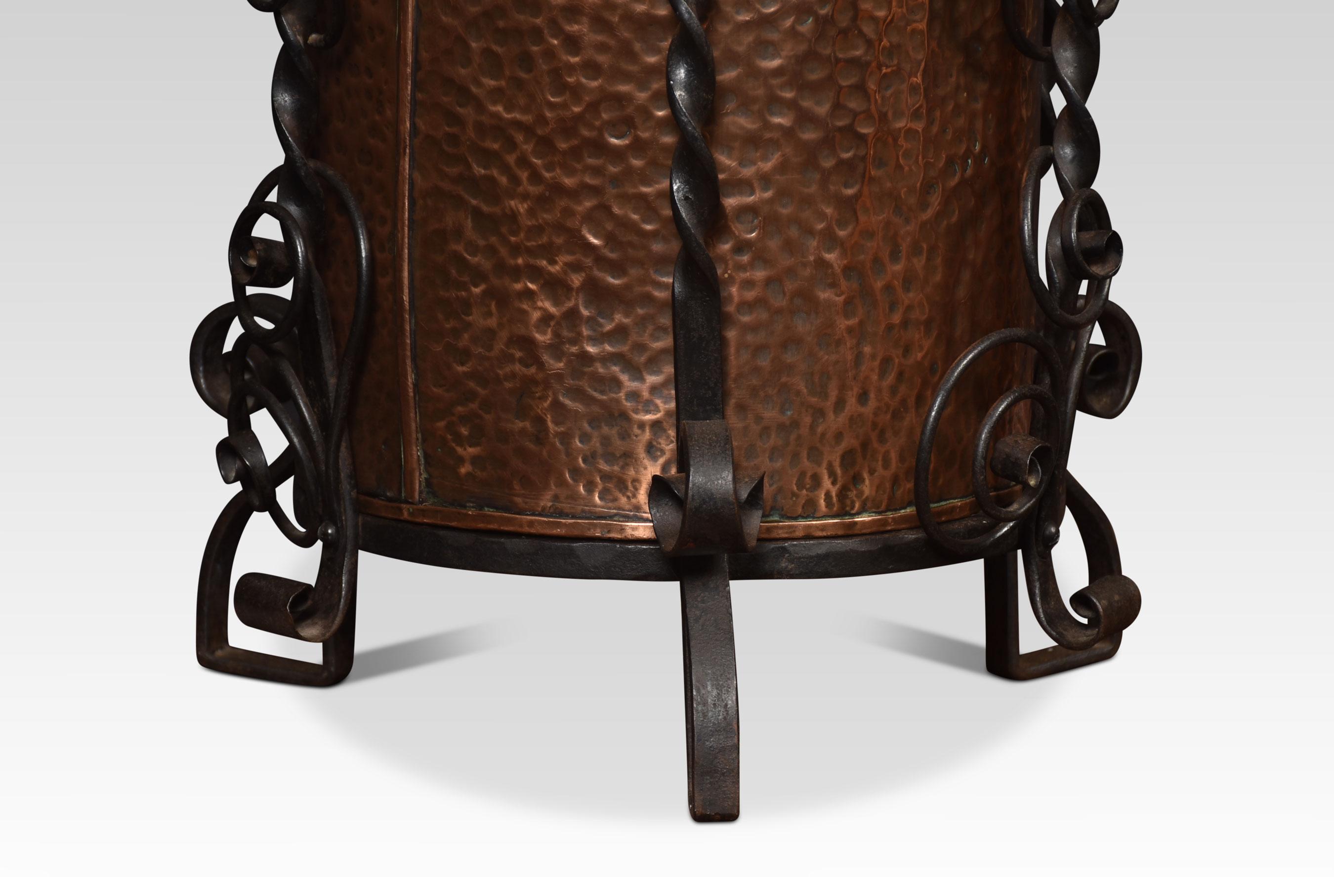 Arts and Crafts hand-planished circular copper coal bucket within a protective black wrought-iron stand, to the dome-topped lid decorated in repoussé style with flowerheads above the cylindrical planished body. All raised up on three shaped