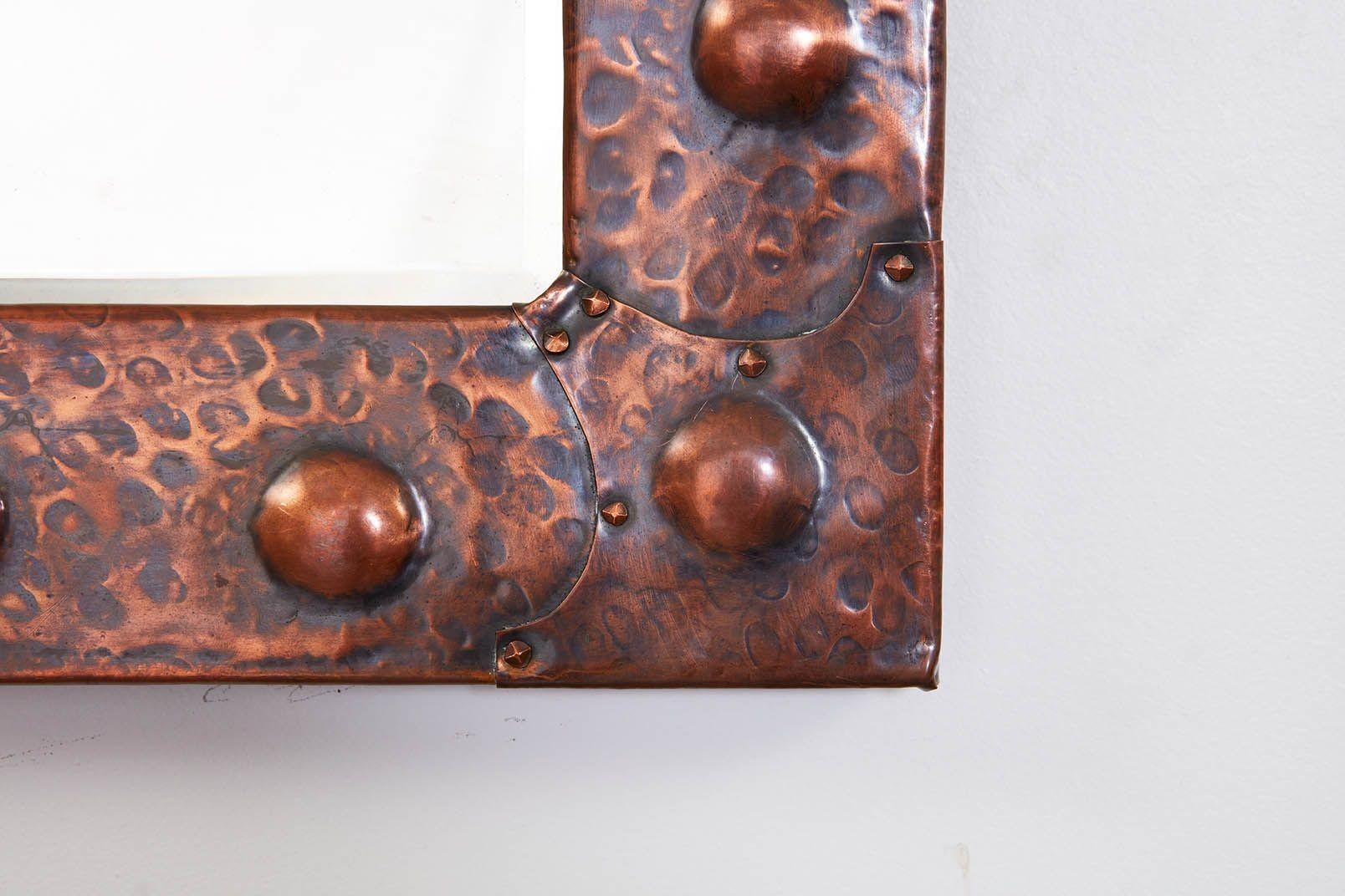 A rectangular Arts and Crafts hand-hammered copper mirror with large copper roundels on a chiseled ground and decorative corner brackets fastened with studs, possibly for Liberty. Retaining original beveled plate glass.