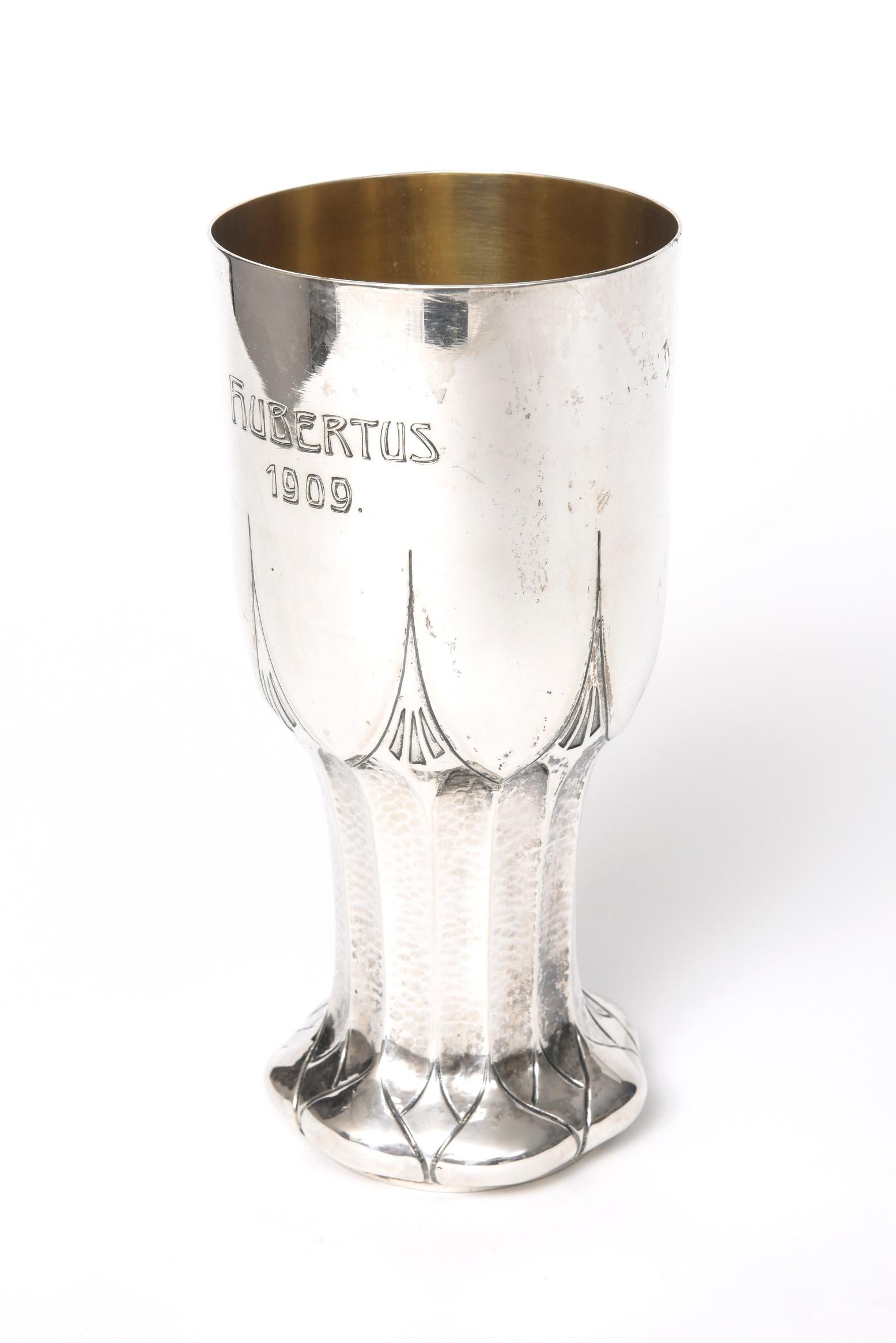 Stupendo calice d'argento 800 di grandi dimensioni, caratterizzato da un gambo martellato con un piede smerlato e punte geometriche che risalgono la coppa. Sulla tazza è inciso Hubertus 1909. Il fondo ha il marchio tedesco 800 e il timbro F. Miller