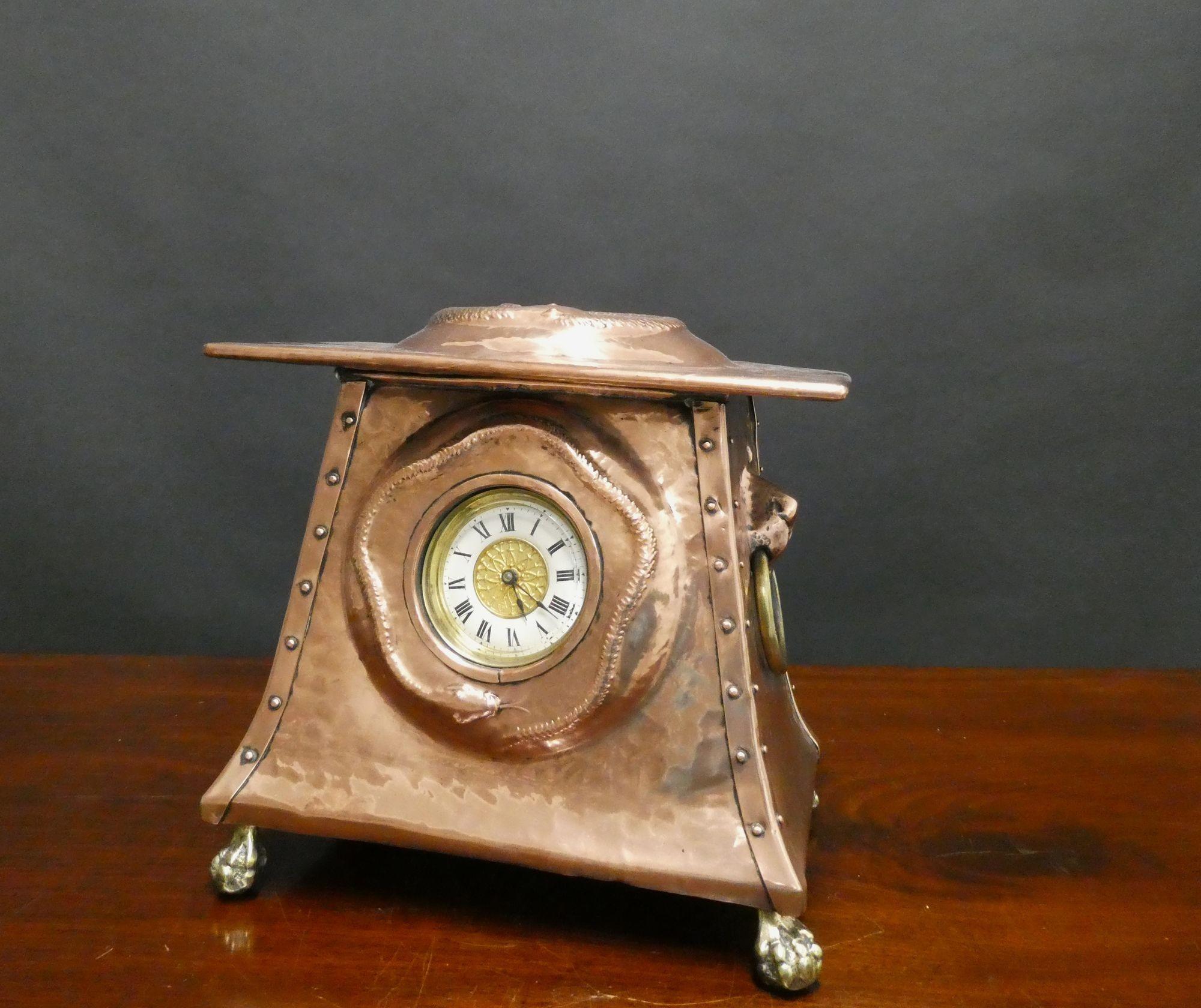 Arts and Crafts Copper Mantel Clock
A most unusual Arts and Crafts mantel clock in an outswept hammered copper case with lions head ring side carrying handles and surmounted by a coiled serpent, resting on four brass lions claw feet.
The dial with