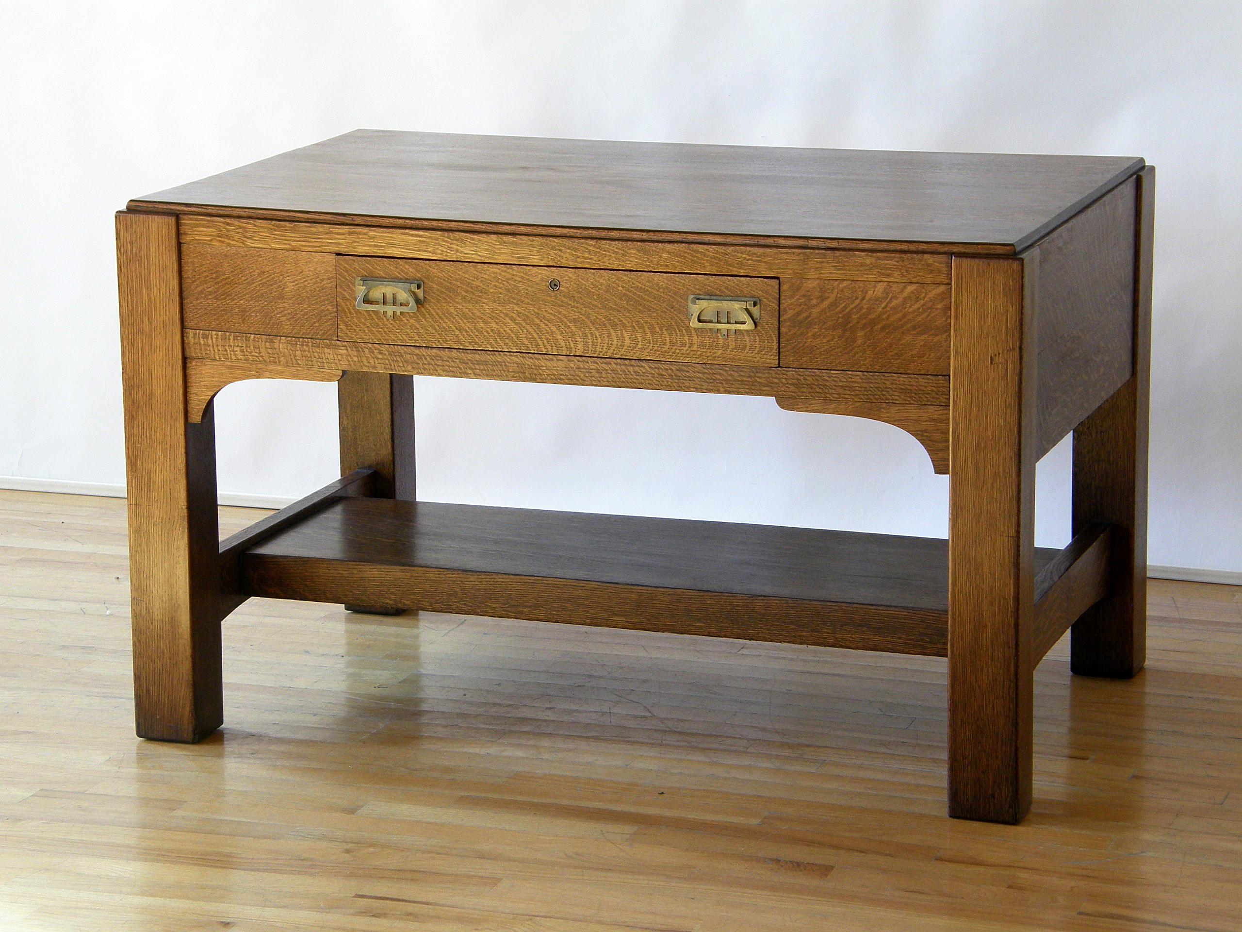 This mission oak library table or desk has a Classic Arts & Crafts style. The drawer has decorative brass pulls and escutcheons with a refined and restrained geometric design. The chunky legs are braced front to back, and those cross pieces support