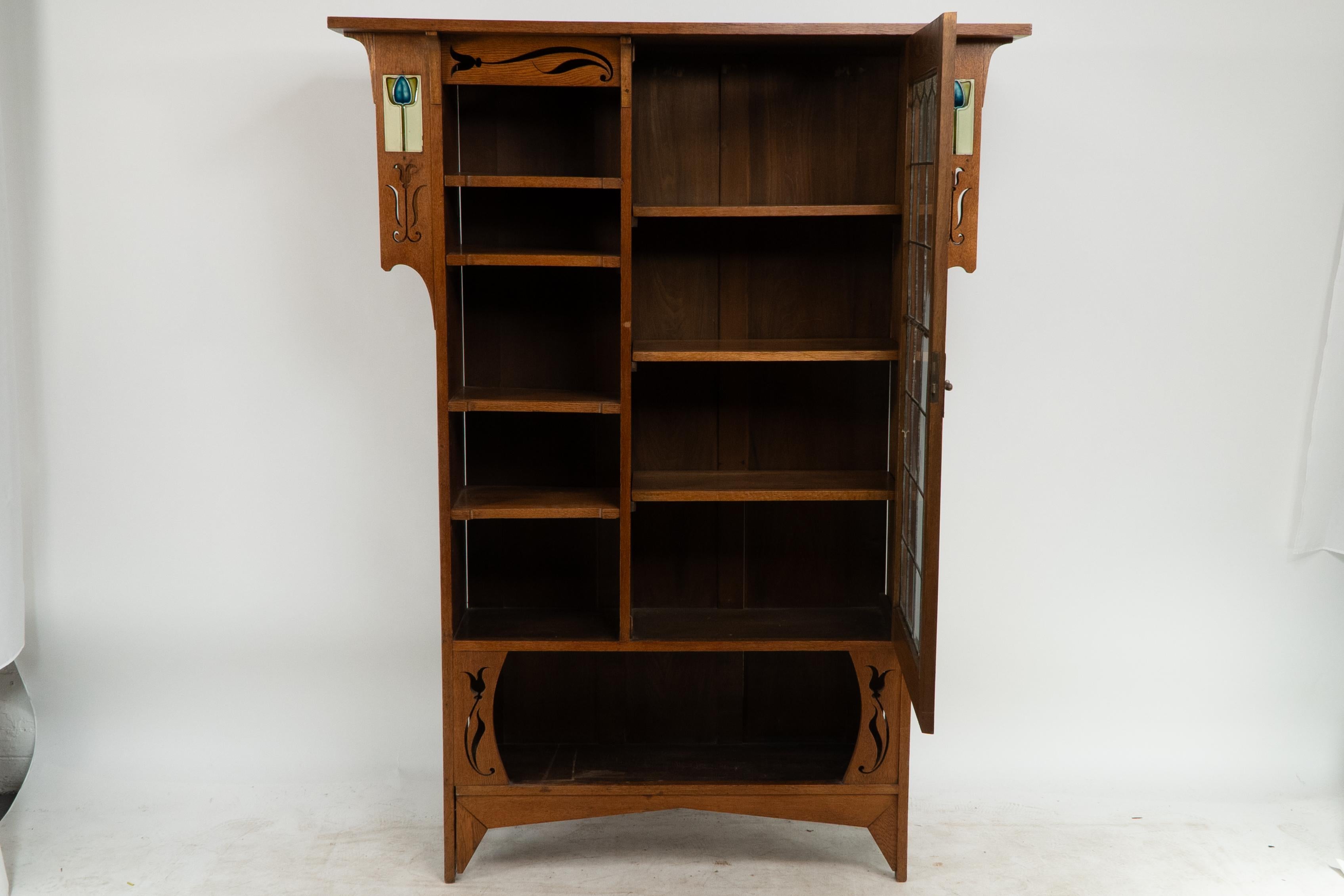 An Arts and Crafts oak bookcase with extended upper sides and inset with original Tulip tiles with stylized floral cut-outs. Adjustable book/display shelves to the left and a stain glass door with decorative hinges and handle.
 