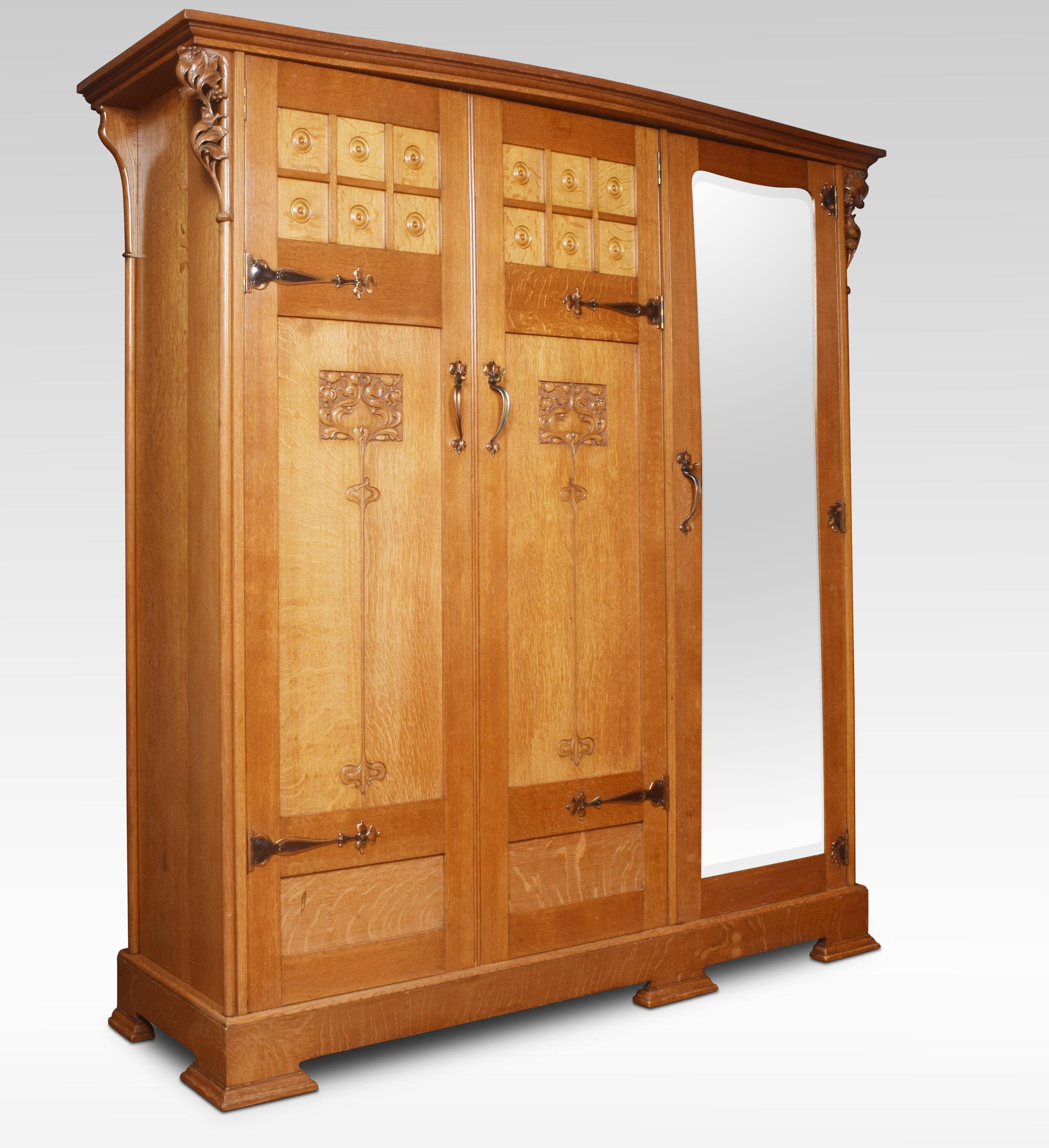 Arts & Crafts Oak Three Door Wardrobe. Having moulded cornice above a large mirrored door flanked with two Panelled Doors opening to reveal large hanging area and drawers to the base. The doors decorated with copper strapwork hinges, tulip carvings