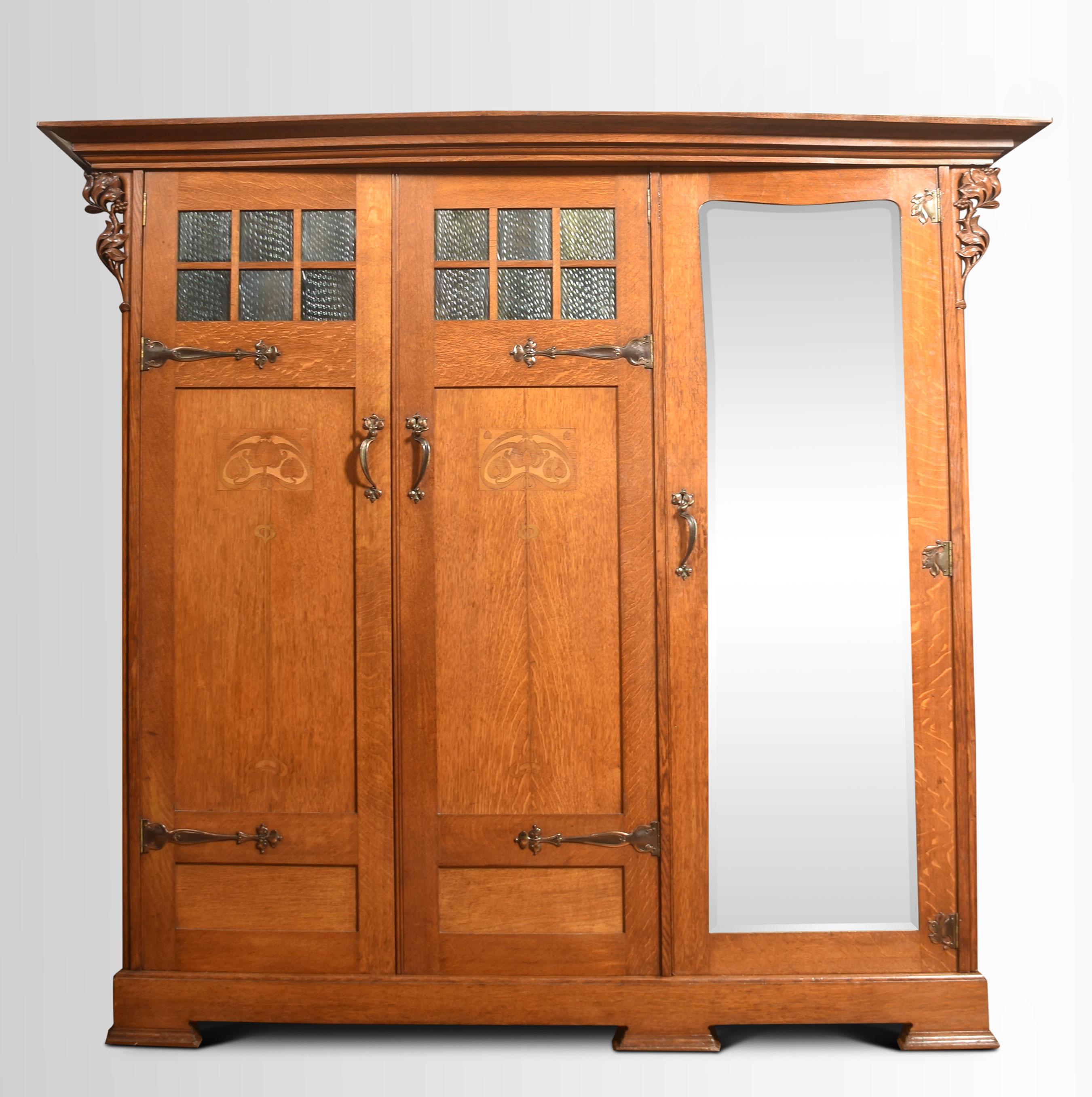 Arts & Crafts Oak Three Door Wardrobe. Having molded cornice above a large mirrored door flanked with two Panelled Doors with unusual glasswork panels opening to reveal large hanging area and drawers to the base. The doors are decorated with copper