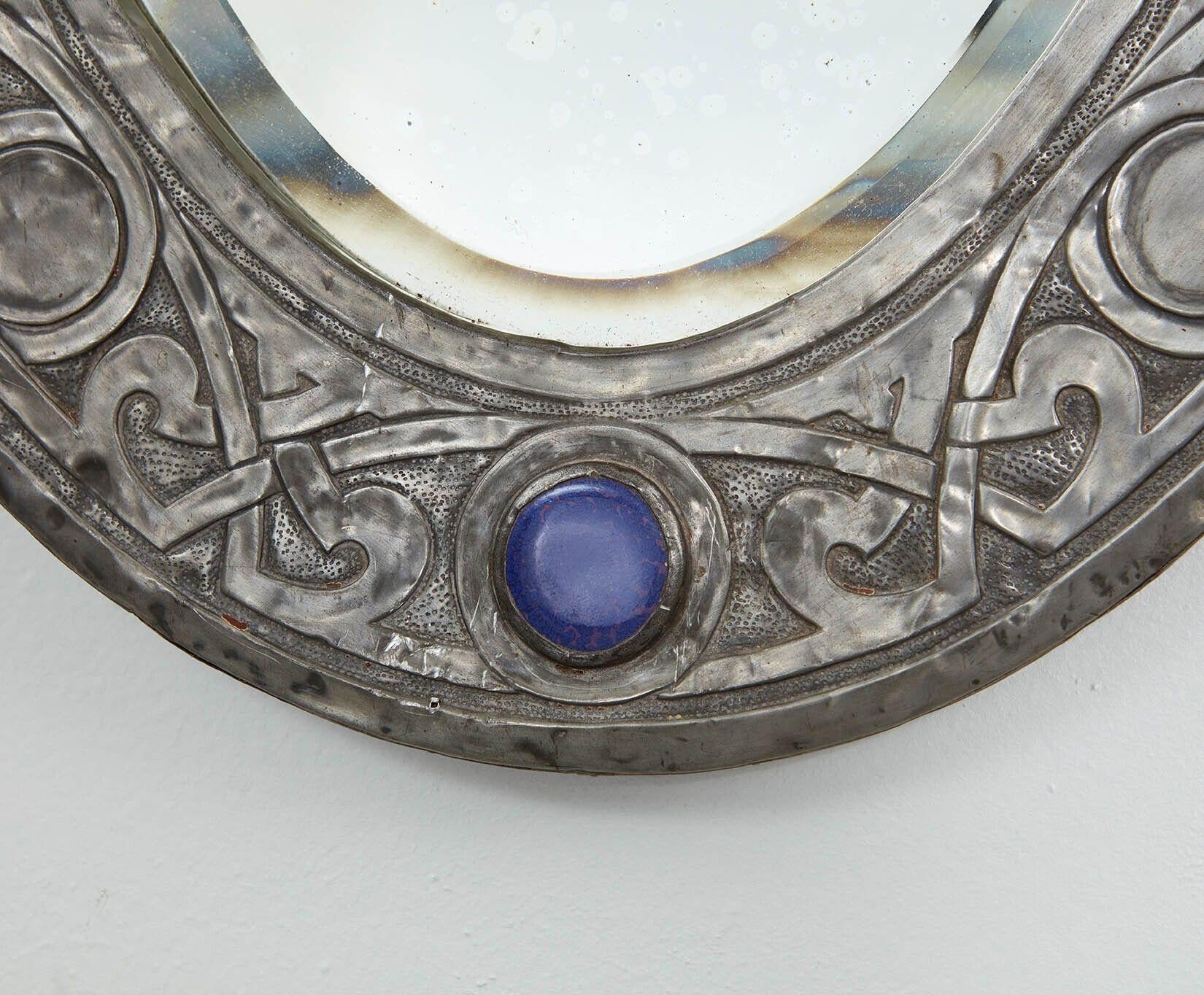 An Arts & Crafts oval mirror in hammered pewter with Celtic strapwork embossing on a punched ground, and having round blue enamel cabochons on the top and bottom, retaining original beveled mirror plate and original backboard.