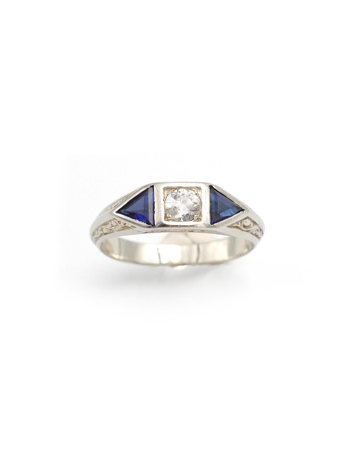 Arts and Crafts ring in 18 karats white gold. Early 20th century ring, centered by a 0.30 ct old-cut diamond, framed by two synthetic sapphires cut into triangles. The band is decorated with vegetal carvings and arabesques. From a men's collection,