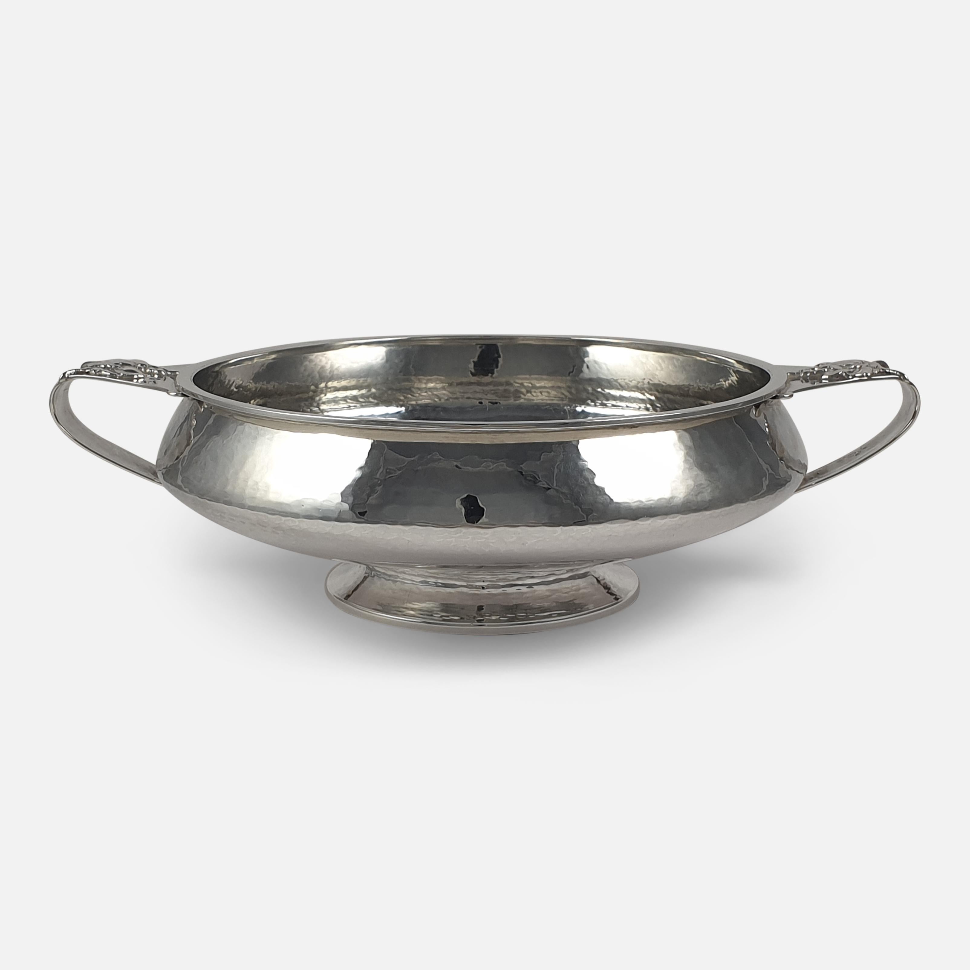 An Arts & Crafts sterling silver twin-handled bowl by A. E. Jones, Birmingham, 1928. The two handled bowl is of circular form, spot-hammered decoration, the side handles applied with grapes and vine leaves, on a raised circular foot.

Assay: -