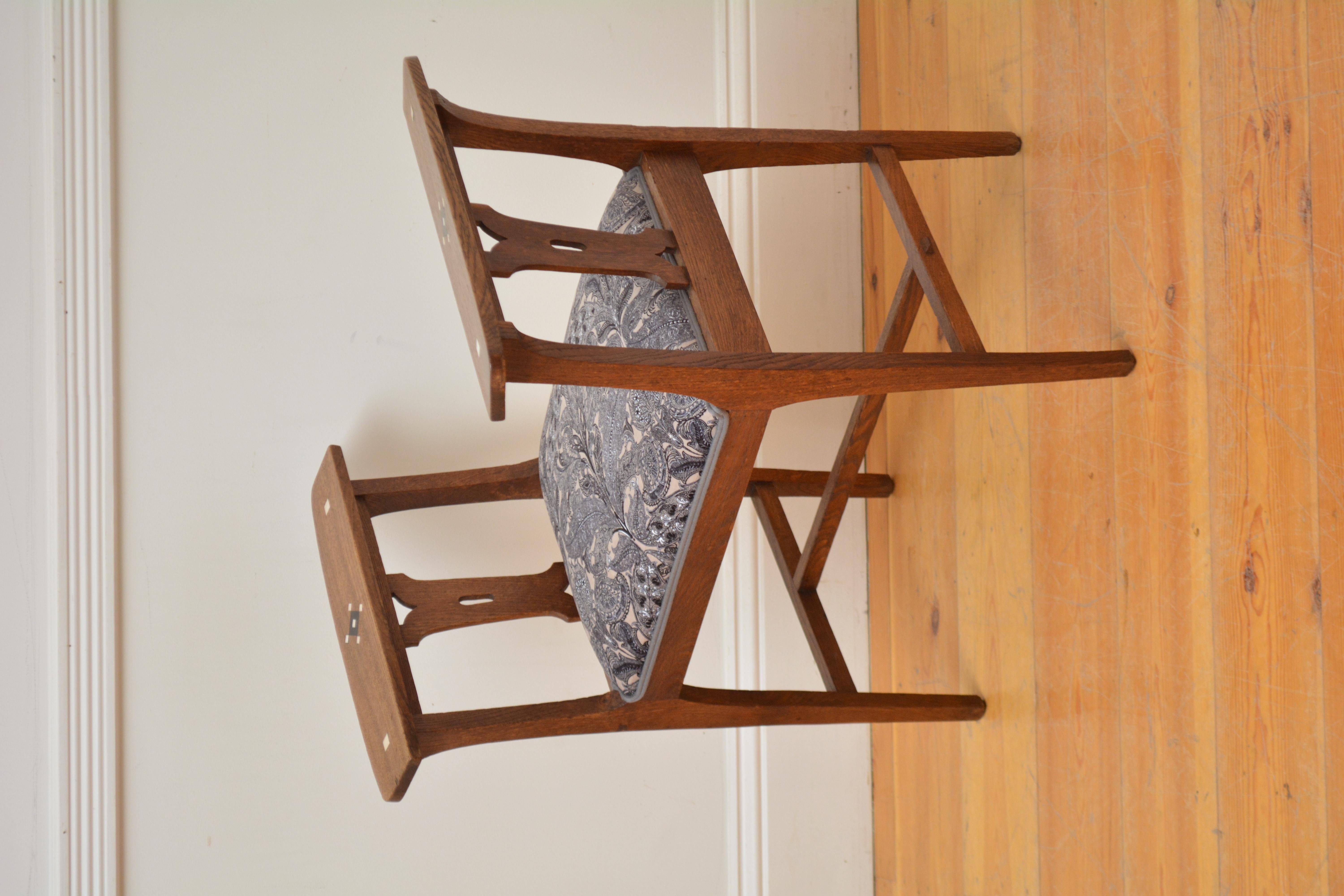 Sn5603 Elegante taburete Arts and Crafts, con asiento recién tapizado en tela floral gris oscuro y beige, flanqueado por brazos con incrustaciones geométricas y soportes tallados, todo ello sobre cuatro patas cuadradas y ahusadas unidas por una