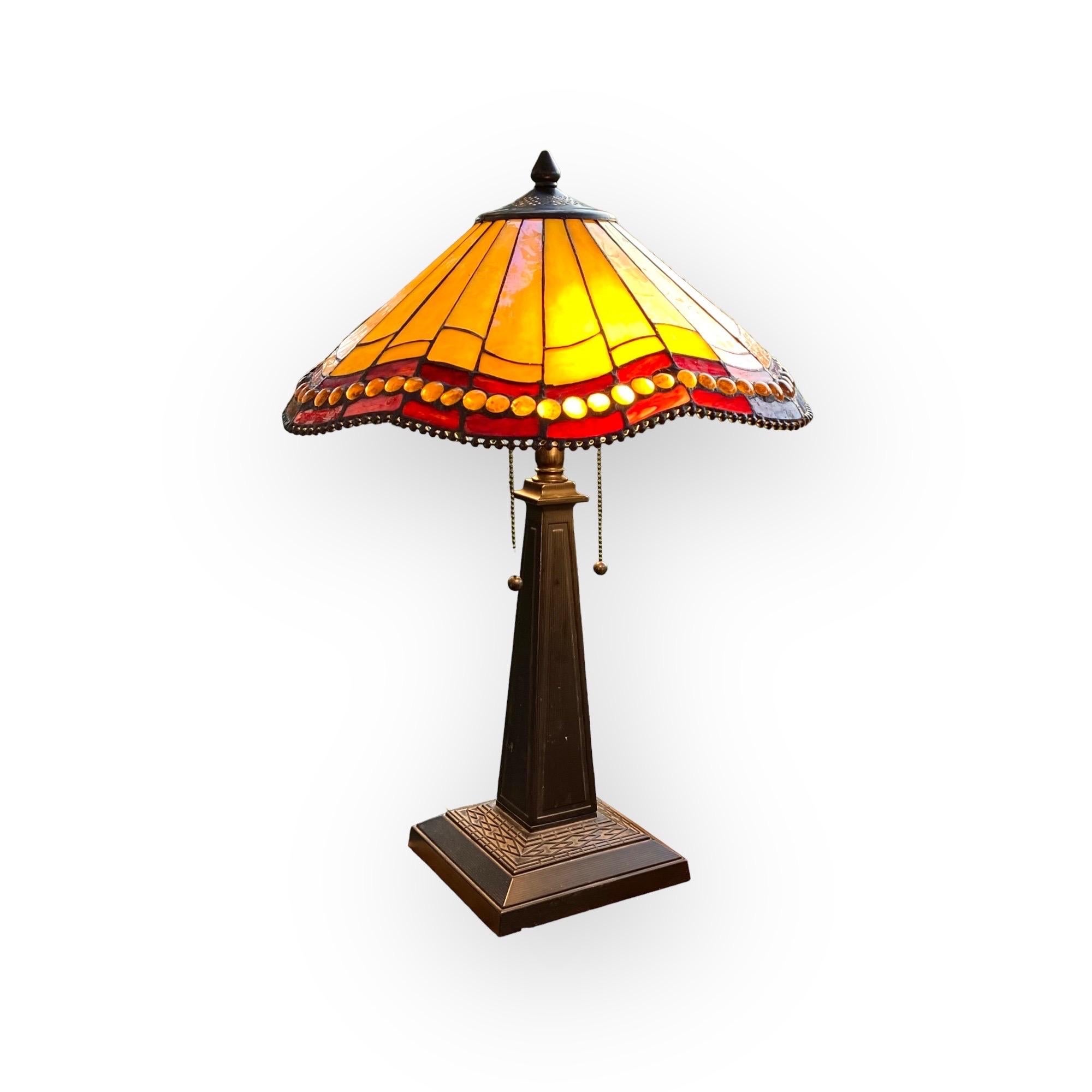 A charming Arts and Crafts style table lamp with a stunning and quality Tiffany style-stained glass shade consisting of slag glass, and stained-glass buttons with bead trim. The heavy, square metal base with two ball and chain pulls for two lights.