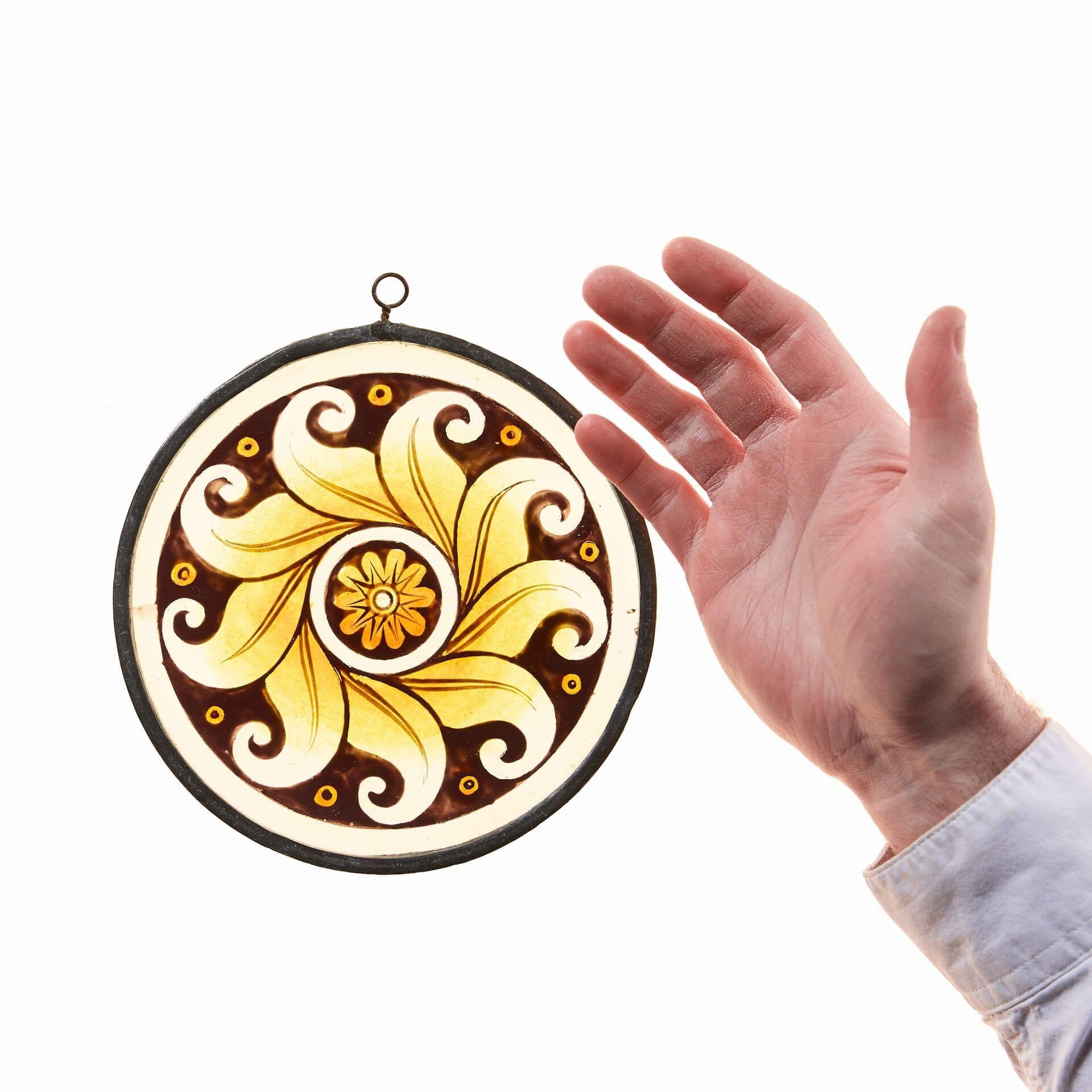 Eine kunstgewerbliche Glasmalerei aus dem späten 19. Jahrhundert, die ein stilisiertes Sonnenmotiv darstellt.

Eine handgemalte, stilisierte, leuchtend gelbe Sonne auf ockerfarbenem Grund, umgeben von einer hellen Umrandung, hebt die lebhafte Farbe