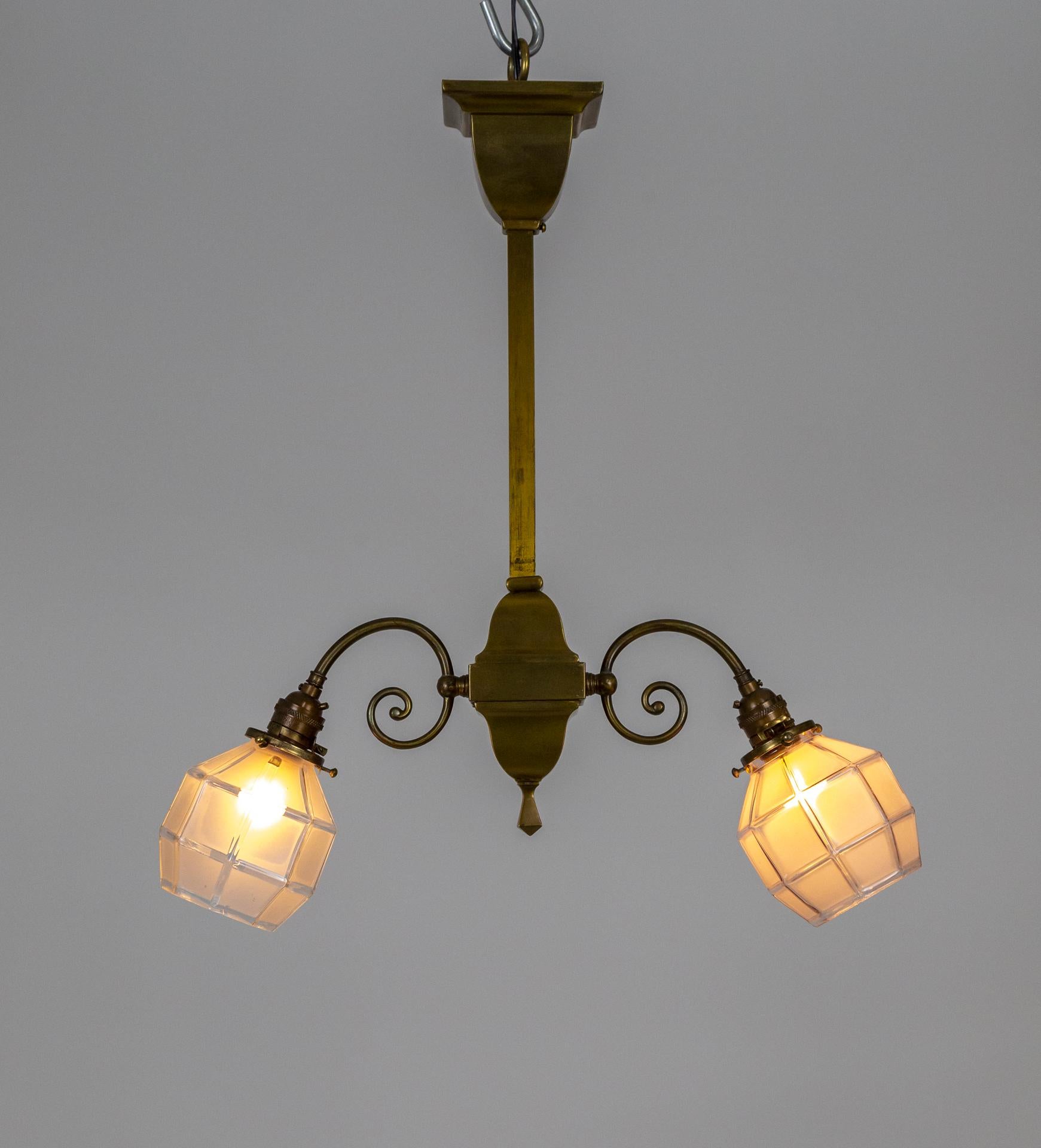 A pair of well-crafted, brass pendant lights with square tube stems and small scroll arms. Added interest comes from the shape of the paneled, frosted glass shades. Early 20th century. Each fixture has two medium bulb sockets, newly wired. 21