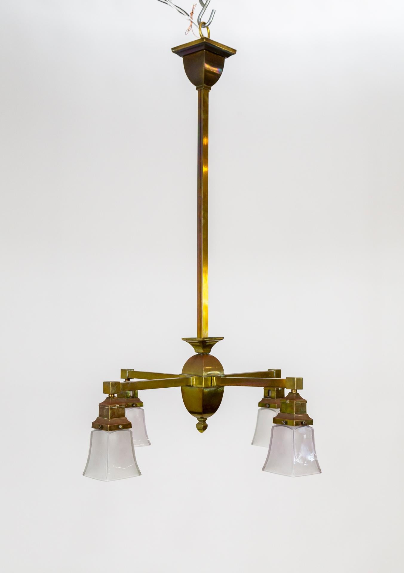 This geometric, brass chandelier is an excellent example of the Arts & Crafts movement. It has a streamlined design with squared stem and arms, and the brass itself has a lovely, patinated finish. The original, glass shades have a glossy exterior