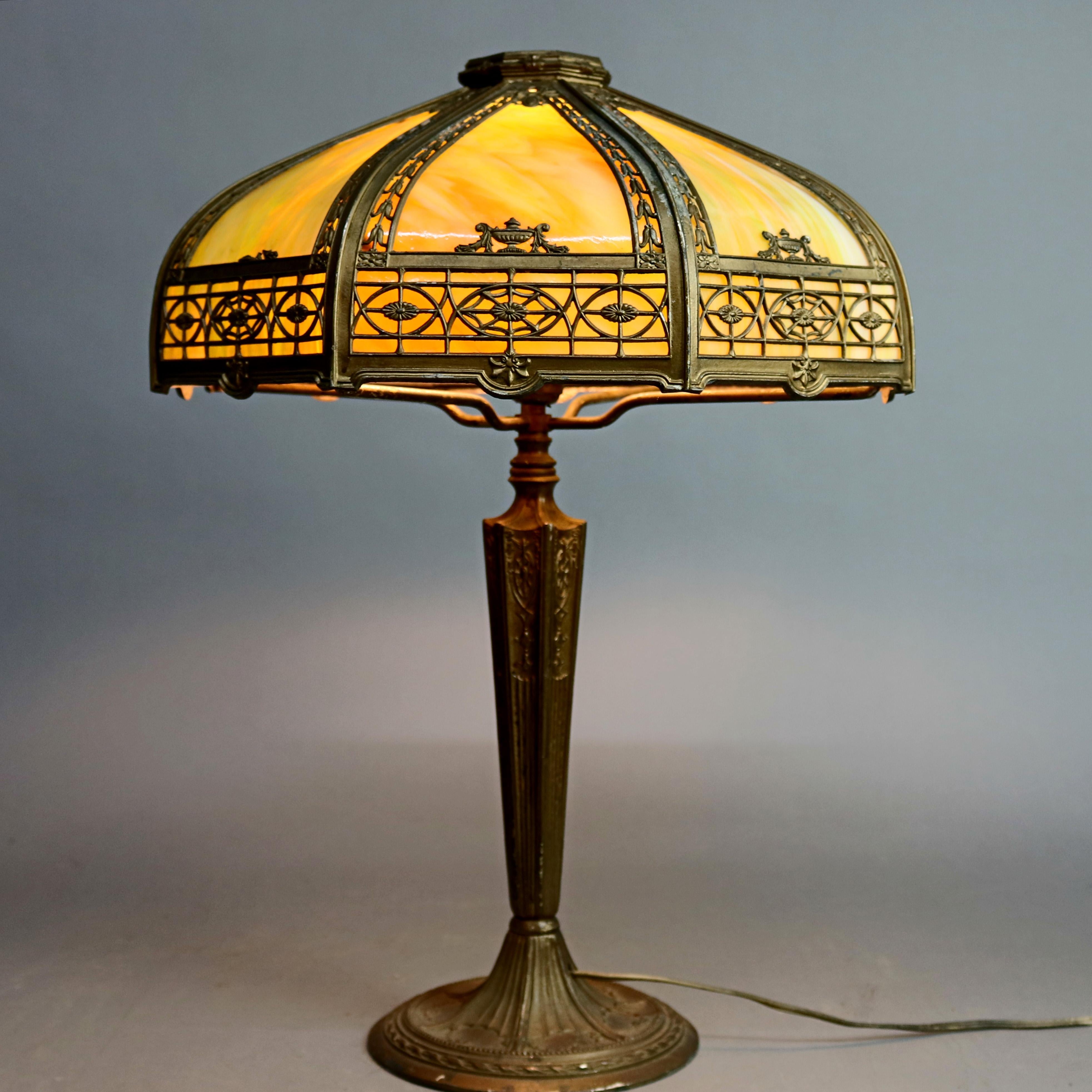 An Arts & Crafts antique Bradley & Hubbard School table lamp offers dome shade with cast and pierced frame having inverted bellflowers and Grecian floral urns housing bent slag glass panels surmounting cast urn form dual socket base with high relief