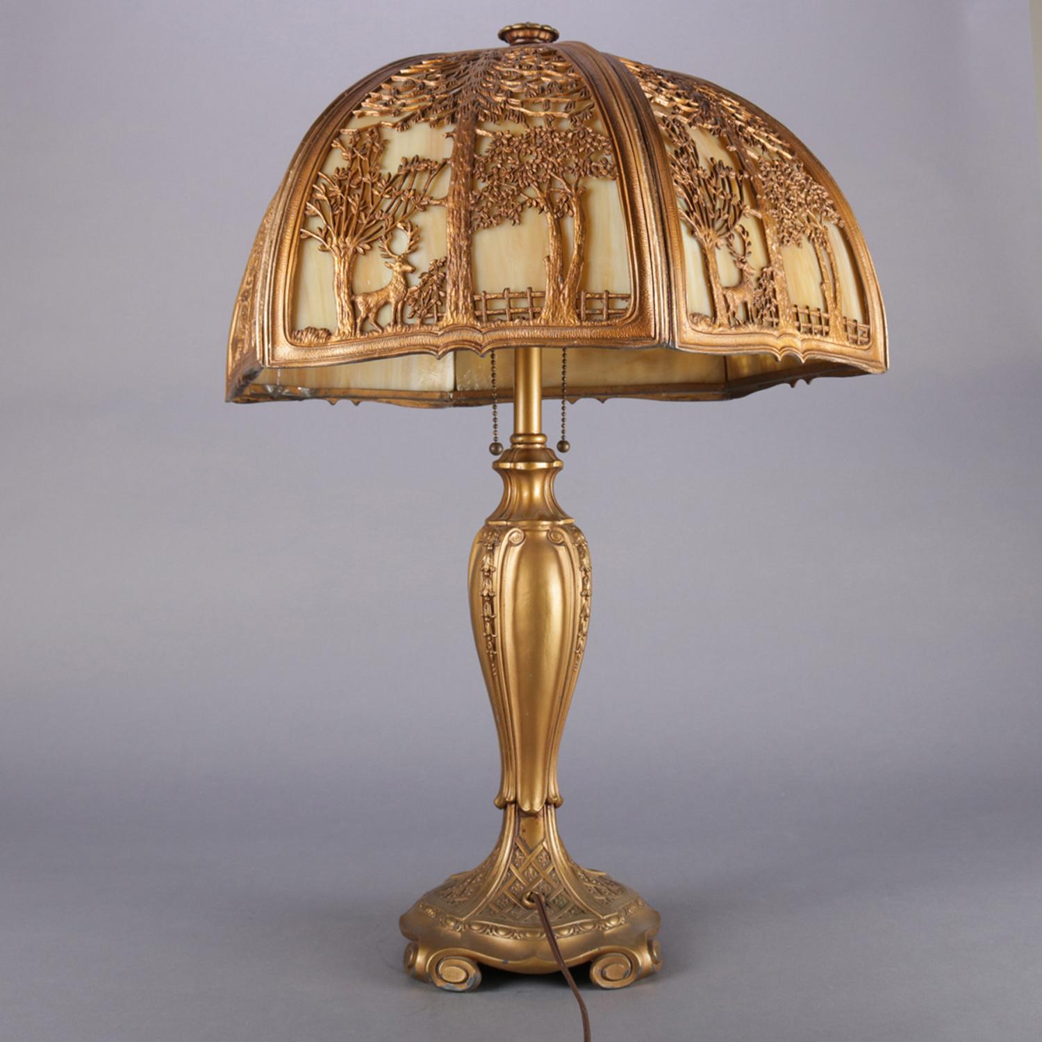 Arts & Crafts Bradley & Hubbard School table lamp features gilt metal base with inverted bellflower decoration and scroll form feet, shade with cast and pierced frame having woodland scene and housing slag glass panels, dual lights with independent