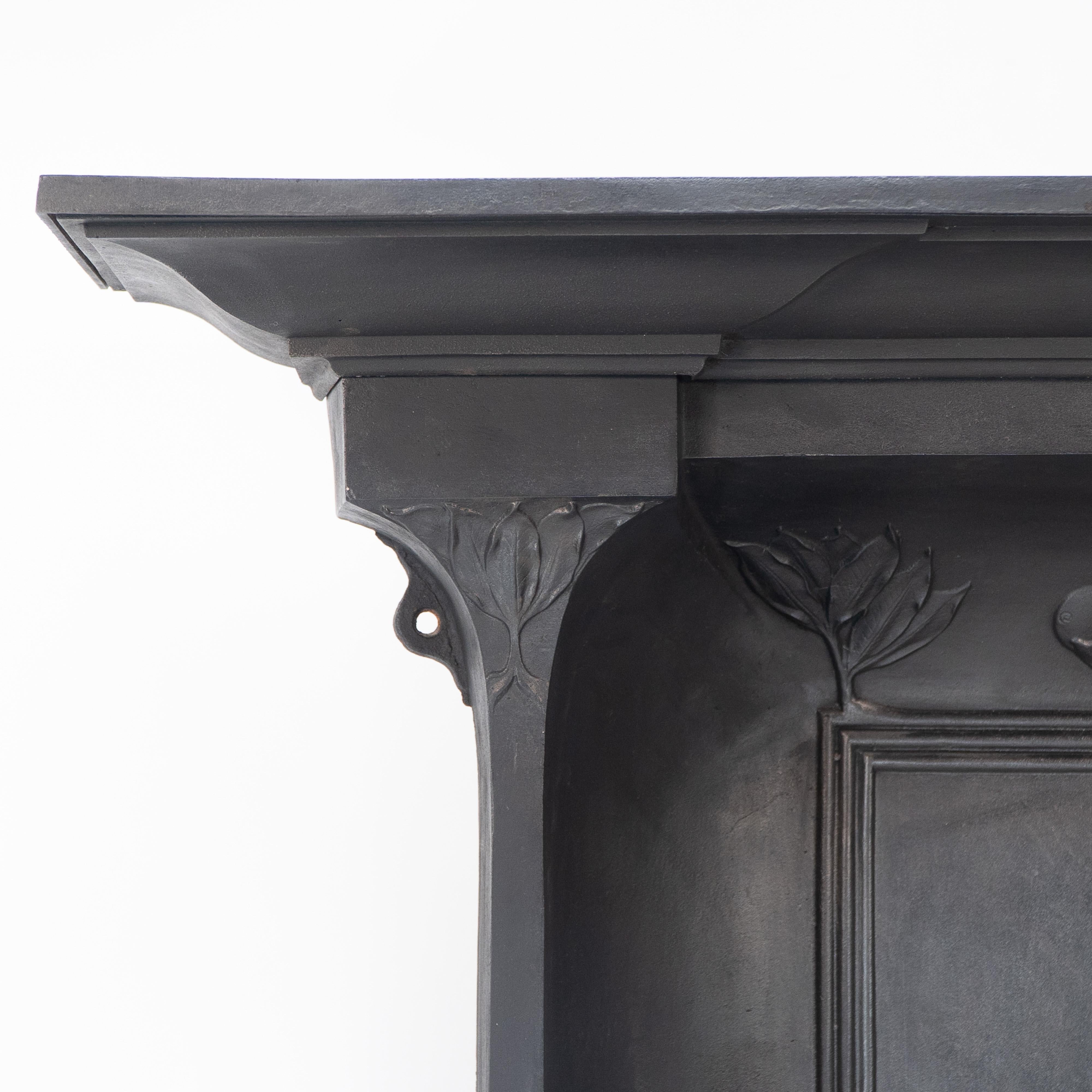 Cheminée en fonte de style Arts and Crafts aux côtés incurvés, conçue pour diffuser plus de chaleur dans la pièce, avec des décorations florales stylisées, des piliers sur les côtés et des étagères chauffantes de chaque côté, près de la base. La