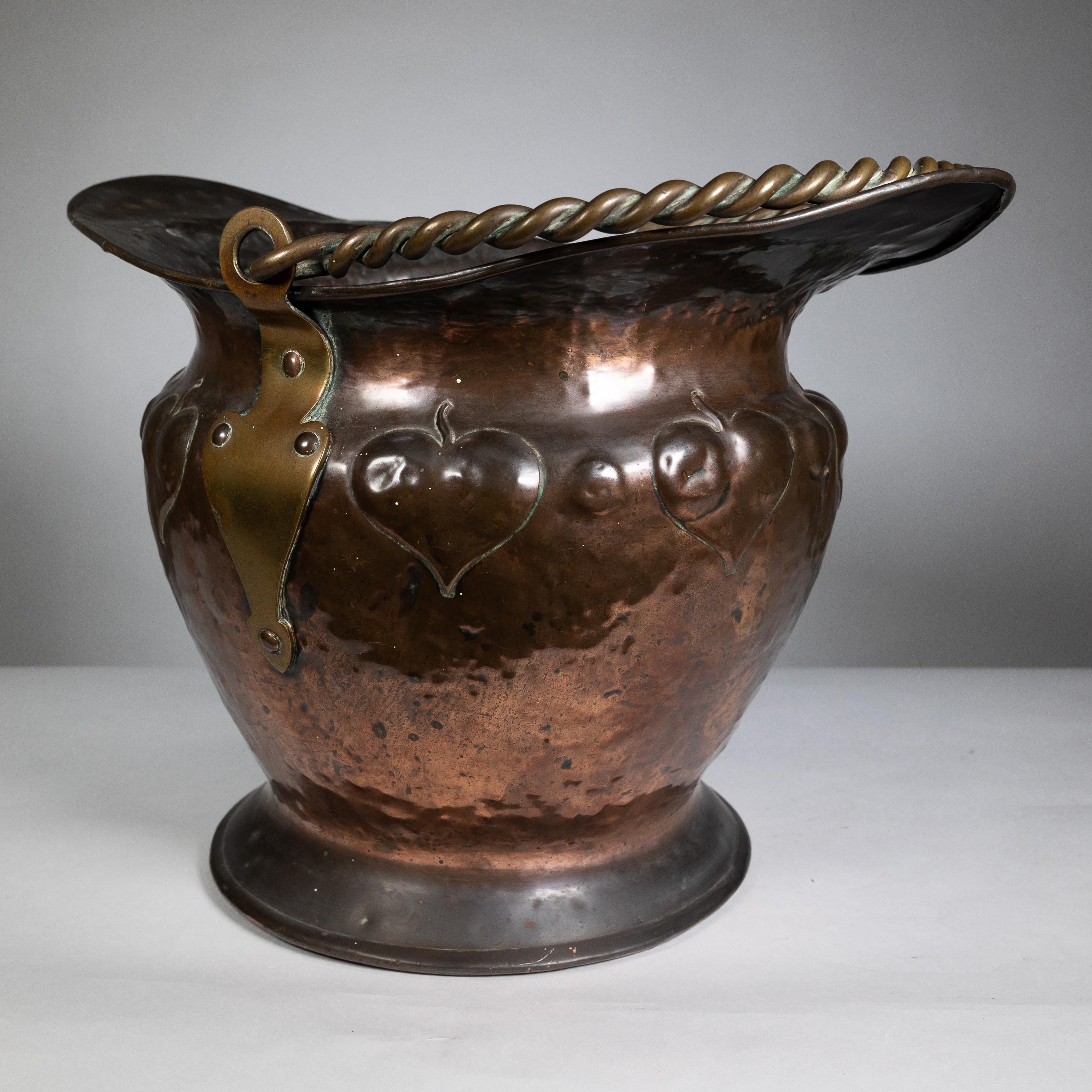Seau à charbon en cuivre de style Arts and Crafts, avec une poignée en corde de laiton et une décoration de feuilles stylisées en repousse autour du corps.