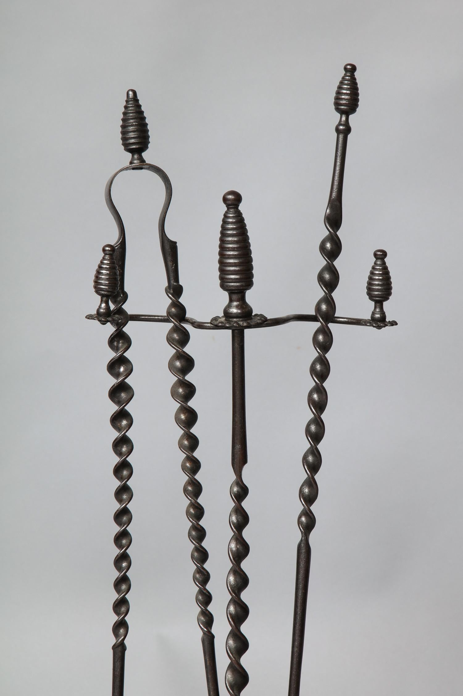 A pair of Arts & Crafts firetools, shovel and tongs, in gunmetal steel with spiral shafts with beehive finials with matching Stand and oval drip tray with raised rim. English, late 19th century.
