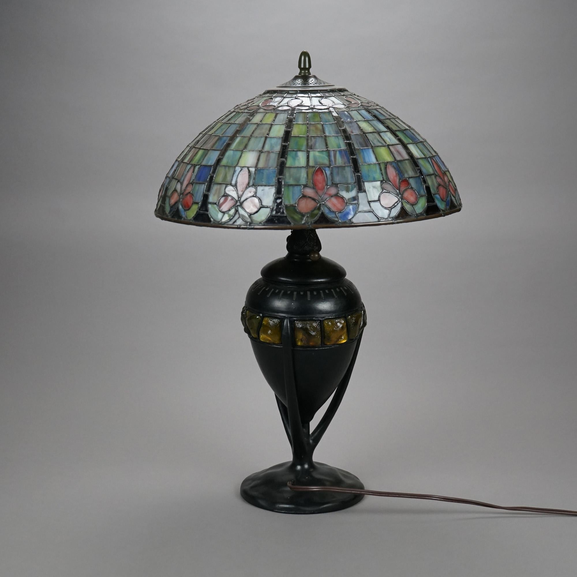 An Arts and Crafts table lamp offers domed leaded glass shade having stylized floral elements over cast urn form double socket base have jeweled art glass inserts, 20th century

Measures- 24.5'' H x 17.25'' W x 17.25'' D.