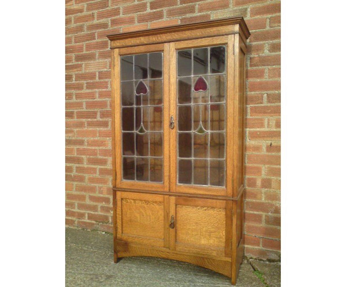 An Arts & Crafts oak bookcase with stylized floral detail to the stained glass doors with a cupboard below, and a sweeping apron to the base.