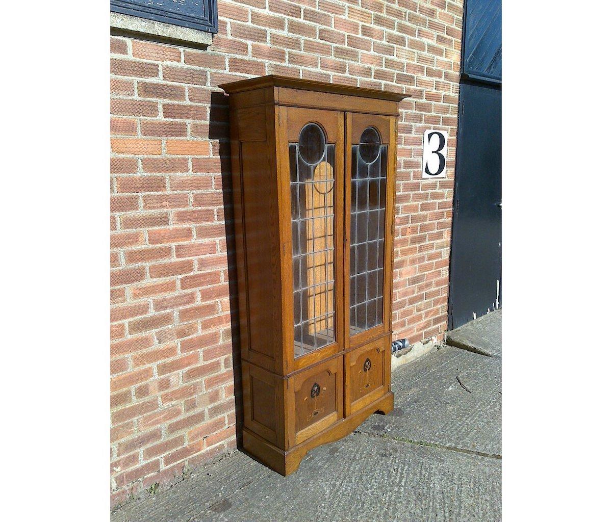 An Arts & Crafts oak bookcase with a flaring cornice, leaded glazed doors with circular details to the tops, the lower cupboard with stylized floral inlays using a combination of pewter, ebonized, and stained sycamore, the whole standing on a double