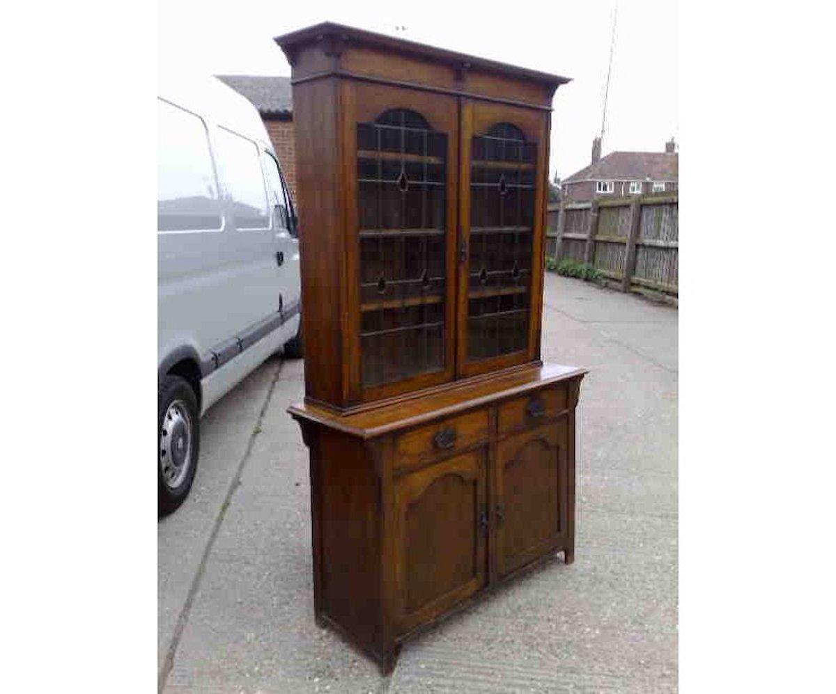 An Arts & Crafts oak bookcase with a flaring cornice and shaped supports below, teardrop stain glass to the leaded doors with arched tops, and a pair of drawers and a cupboard below, also with arched top panels.
Measures: Height 79