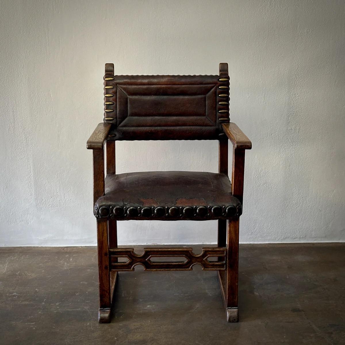This 19th-century English armchair combines historical charm with timeless design. Crafted from oak, it features a decoratively carved front stretcher and richly patinated leather upholstery adorned with large brass studs, adding an air of classic
