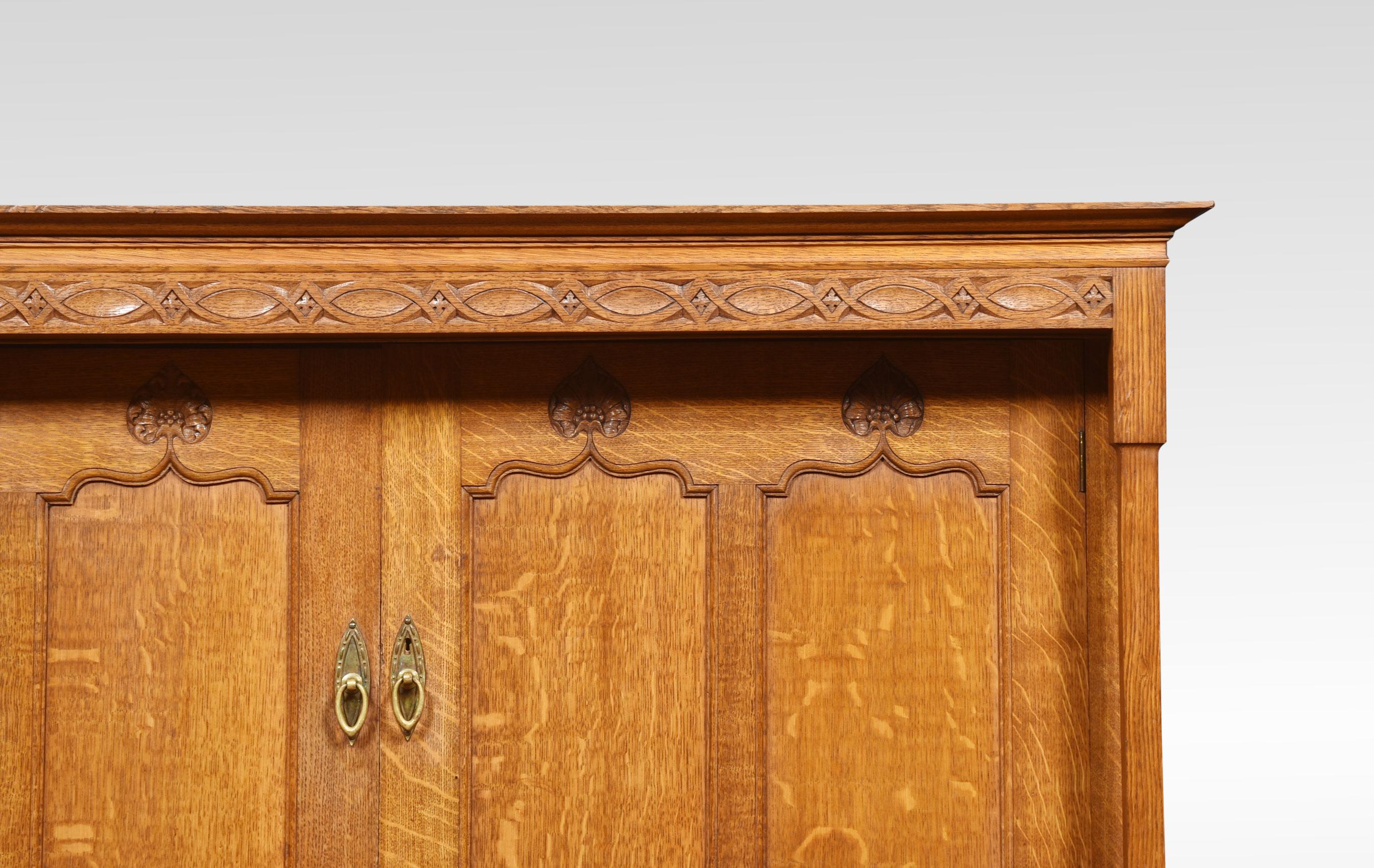 Arts & Crafts oak cupboard the moulded pediment and carved frieze above a pair of panelled doors with lily motifs enclosing a sliding tray, flanked by square tapered columns. Above a projecting serpentine front with four short and two long drawers