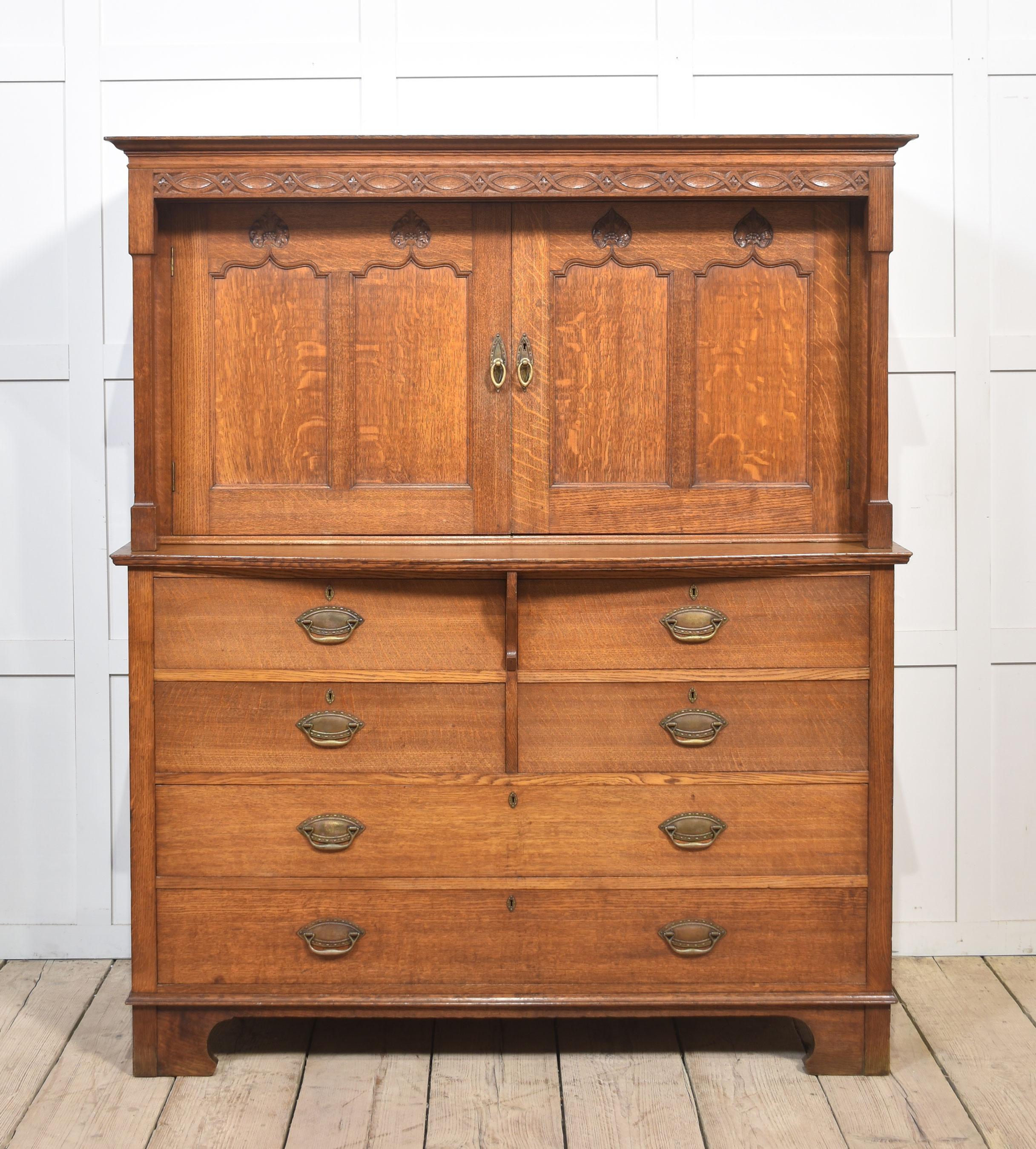 Armoire en chêne de style Arts & Crafts, avec fronton mouluré et frise sculptée au-dessus d'une paire de portes à panneaux avec motifs de lys renfermant un plateau coulissant, flanquées de colonnes carrées et effilées. Au-dessus d'une façade