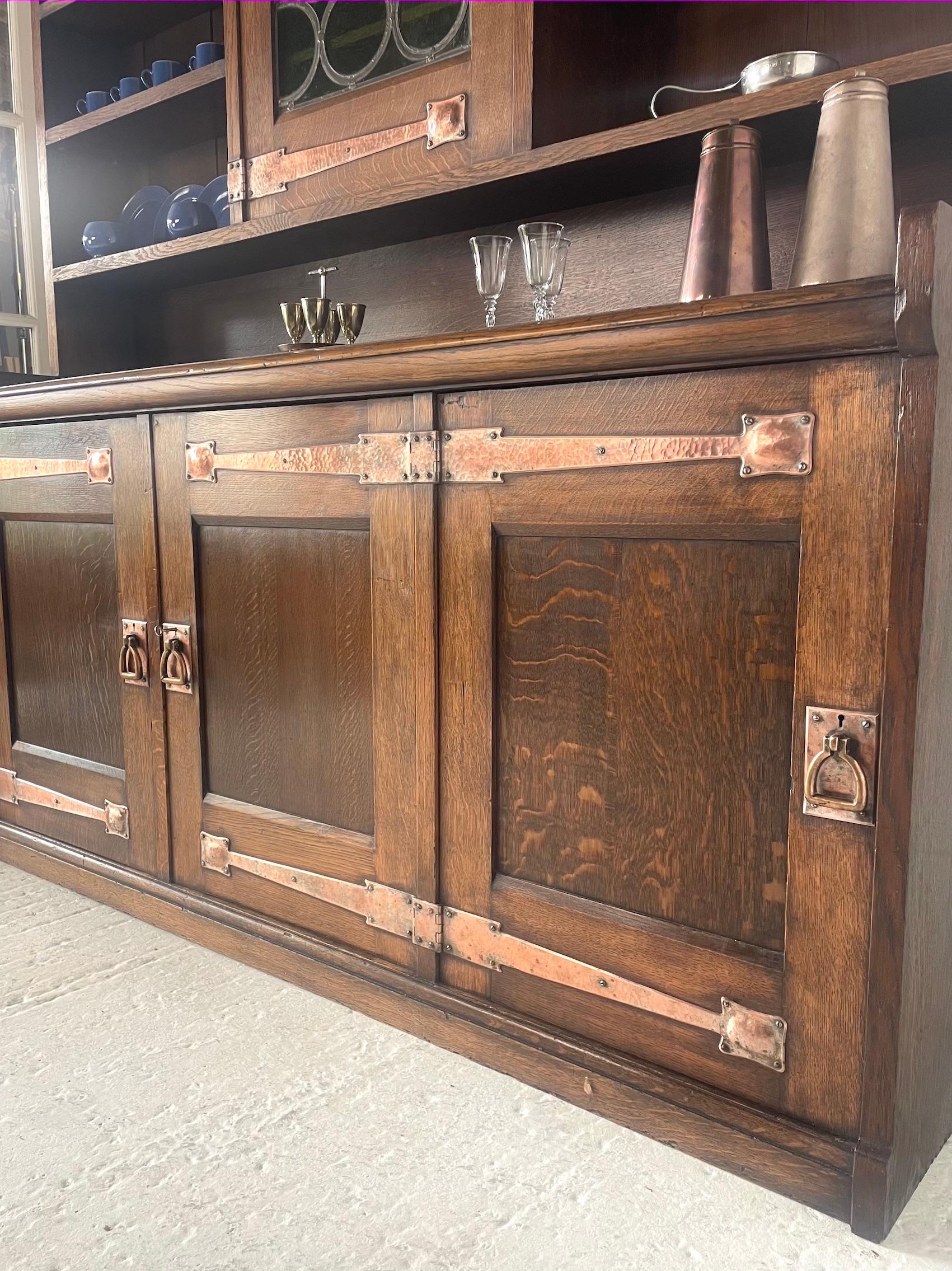 Arts & Crafts Dresser in quarter sawn oak
Stylised copper strap hinges
Copper stirrup handles
Leaded textured green glass panel to the central door
Three doors below
Adjustable shelving
Attributed to the studio of Leonard Wyburd
Liberty & Co
Circa