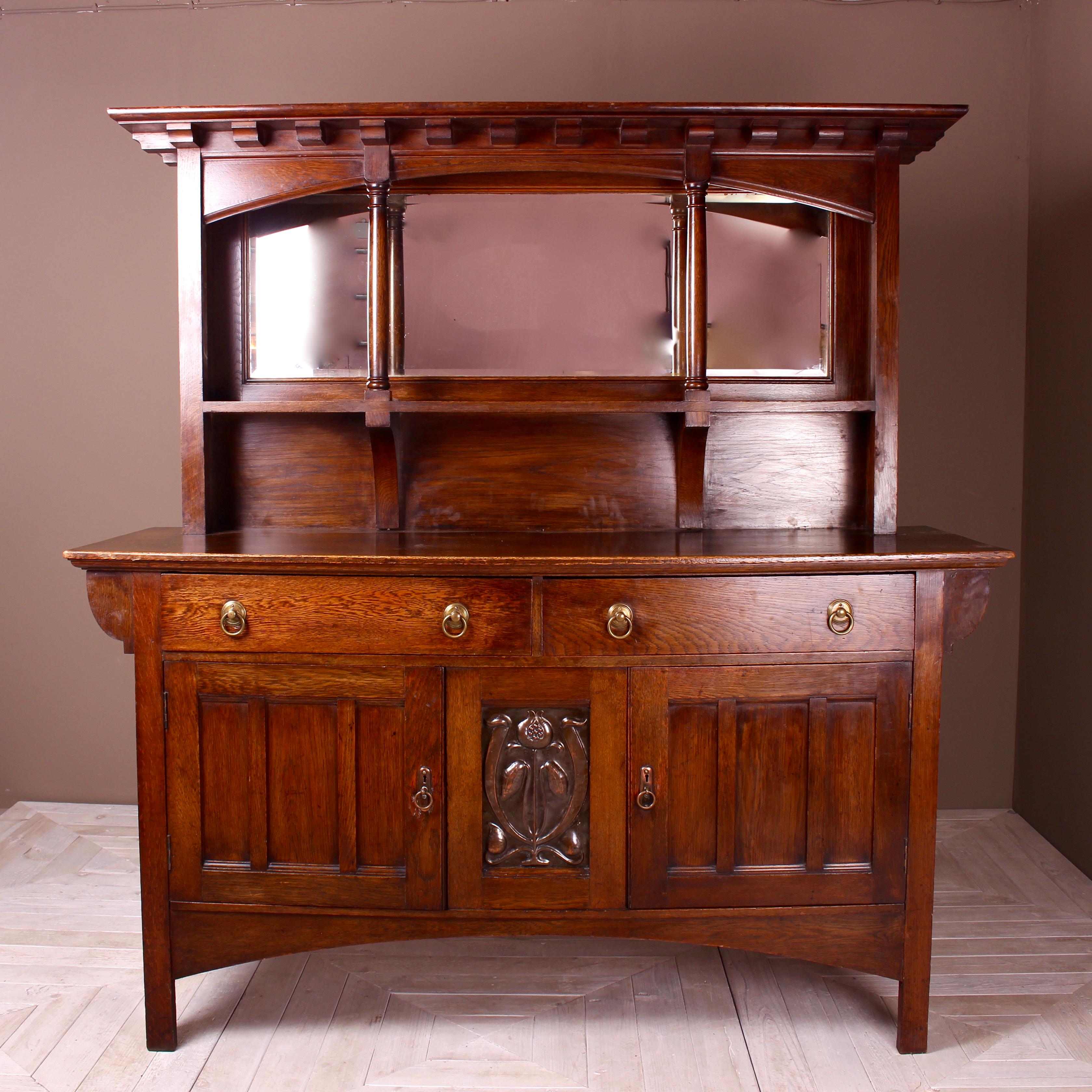 An Arts & Crafts oak sideboard, possibly by Shapland & Petter, the flared dentil cornice above turned supports backed by beveled mirror panel, base fitted two drawers with brass handles, a pair of panel doors below flanking an embossed patinated