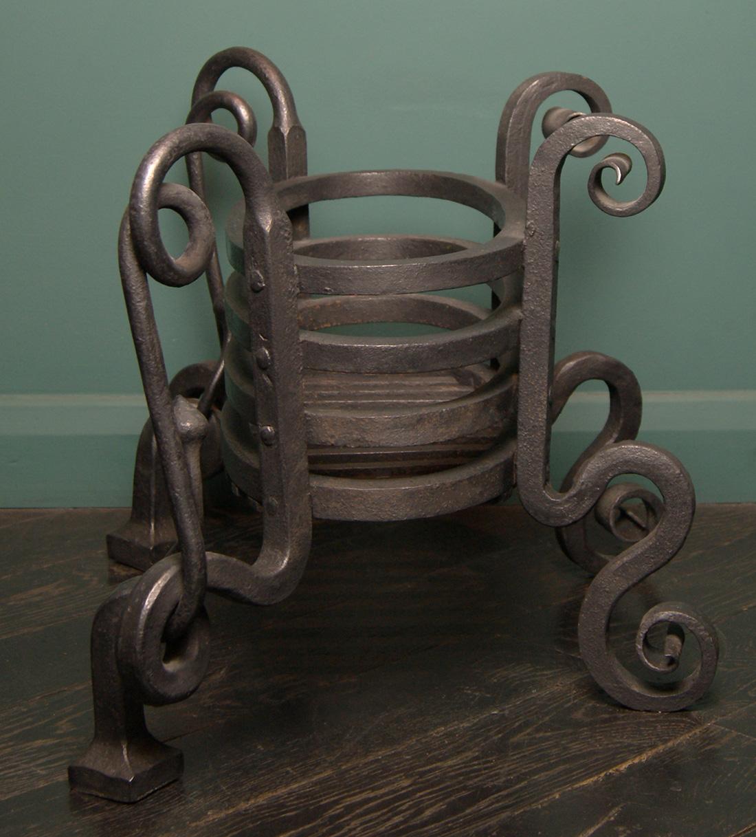 An Arts and Crafts oval wrought-iron fireplace basket. A black wrought-iron railed basket with shaped legs on plinths. The standards extend and scroll intertwining with the legs with turned finials. Restored. Circa early twentieth century.
