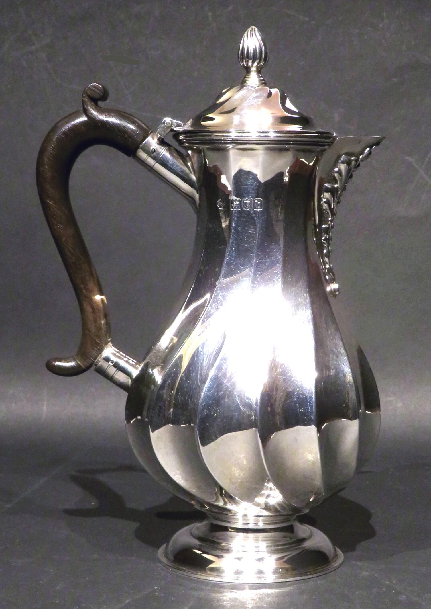 A stunning Arts & Crafts Period sterling silver hot water / hot milk pot showing a sculptural, baluster shaped body expertly worked with swirling fluted panels, rising to a conforming hinged top above a spout with embossed organic decoration, sided