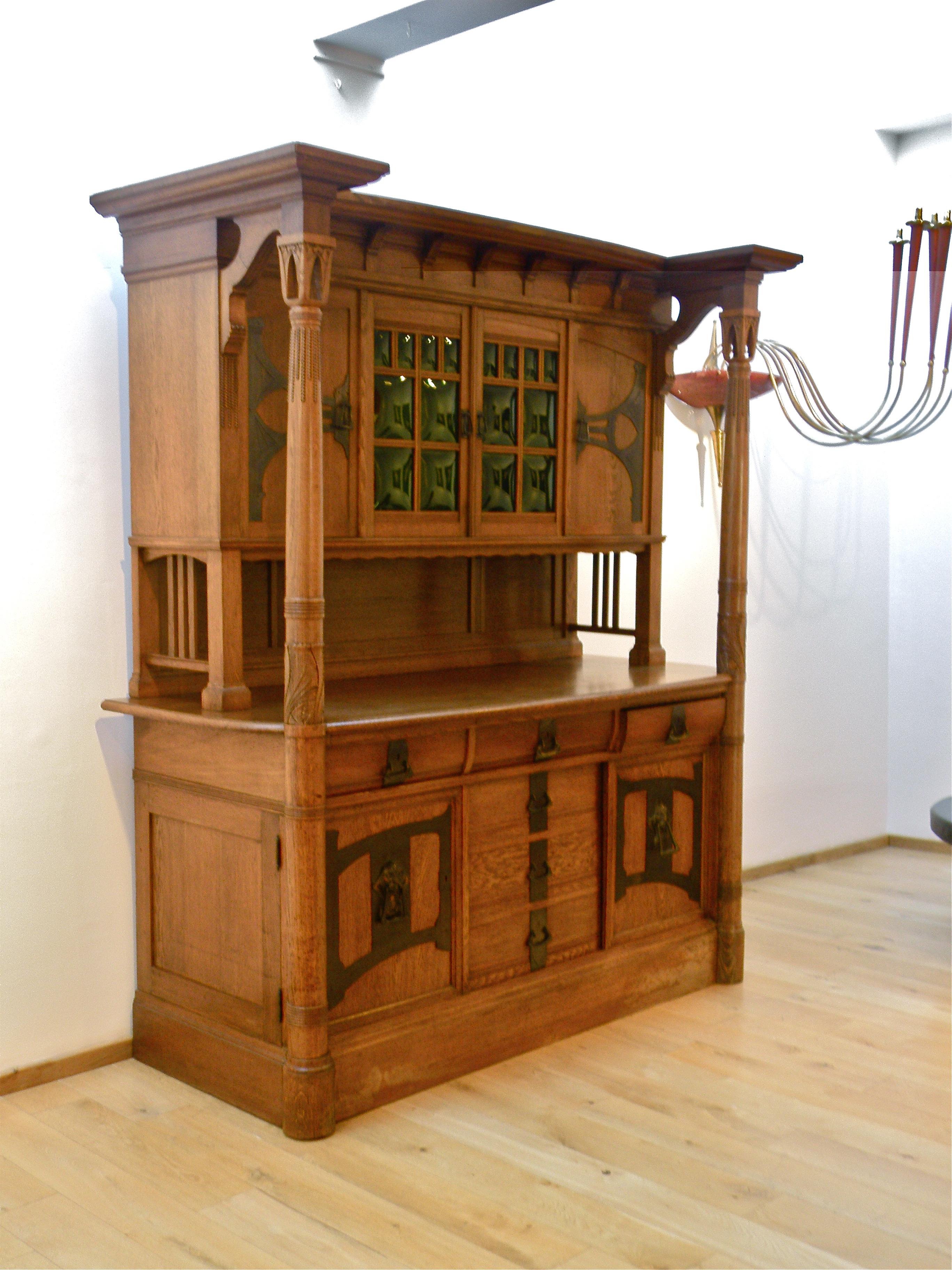 An impressive Northern European Arts & Crafts sideboard.
Oak, with carved stylised lotus blossom capped support columns, green glass panes, brass mounts and drawer and door pulls. (In two sections so can be more easily transported).