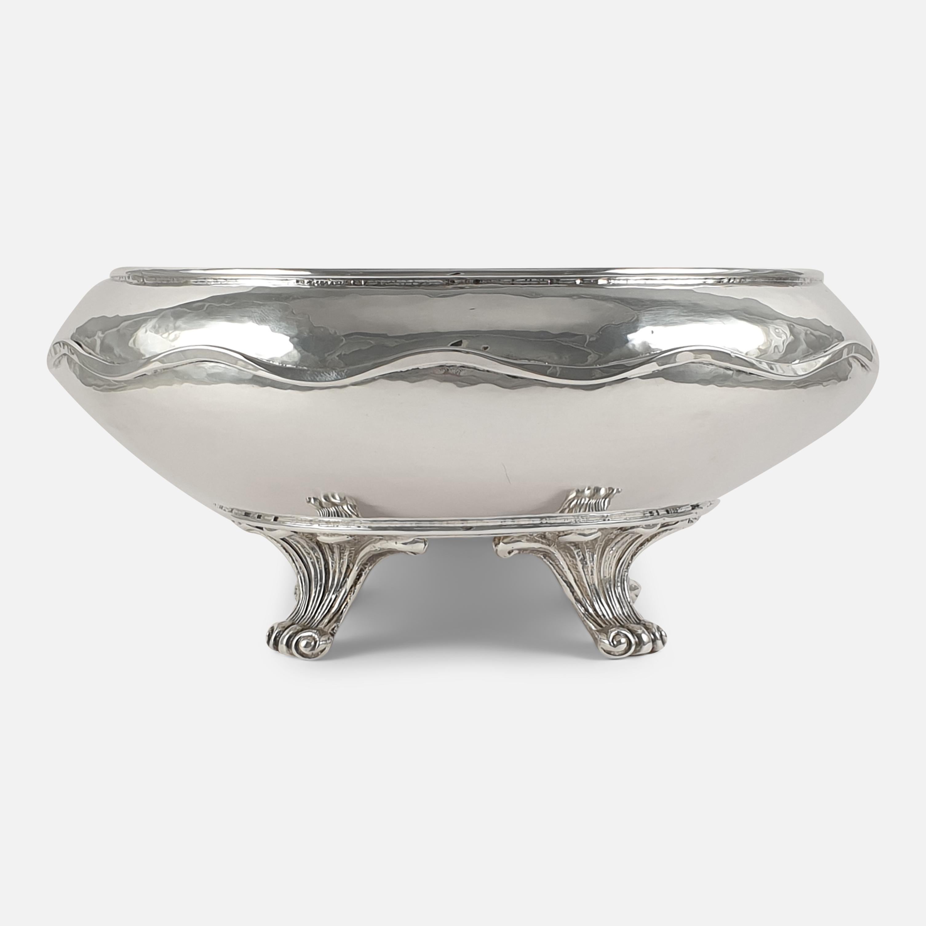 An Arts & Crafts style sterling silver bowl made by Omar Ramsden. The bowl is of circular tapering form, with spot-hammered decoration, and a wavy-edge border, sitting on four fluted scroll feet. 

Assay: - .925 Silver.

Date: - London,