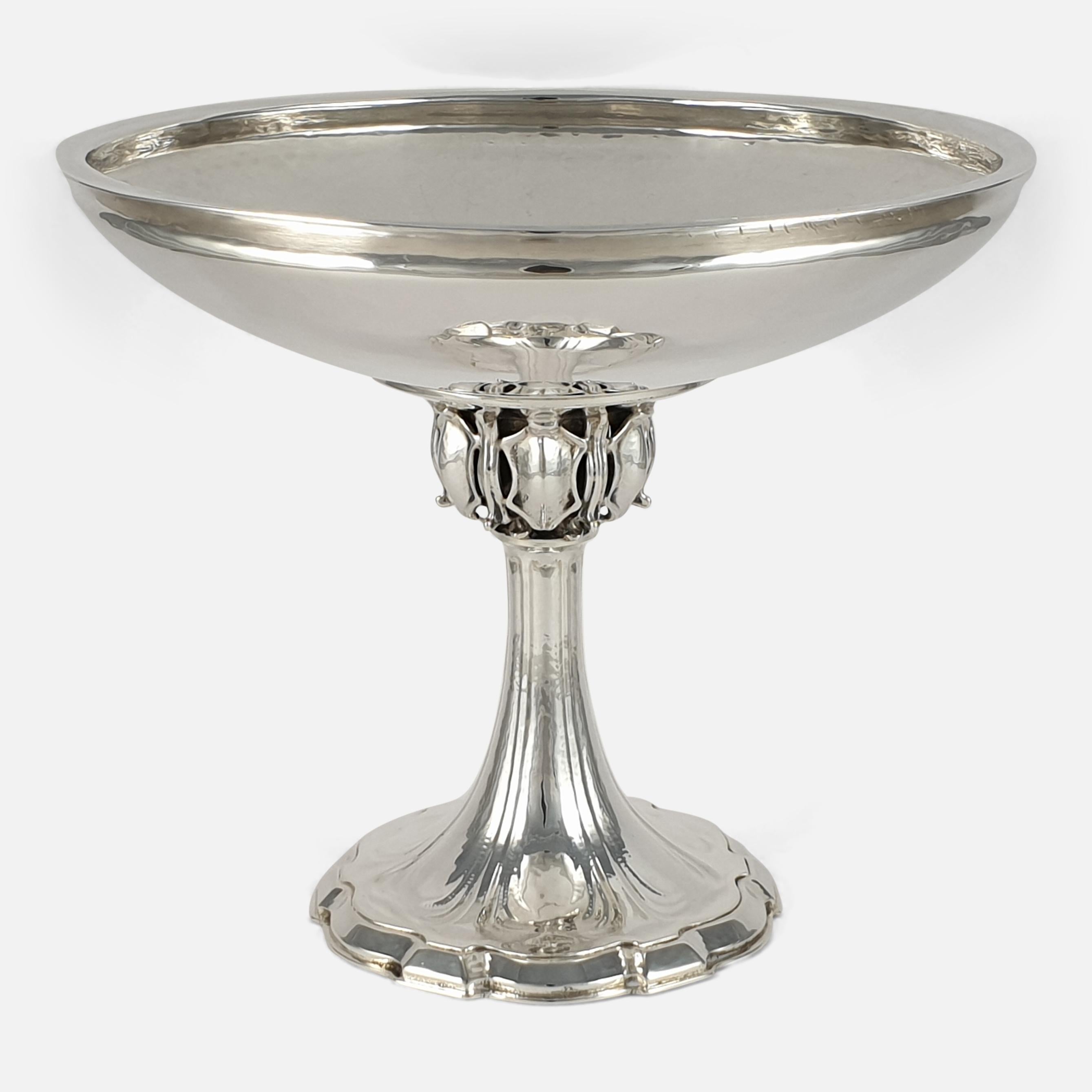 An Arts & Crafts style sterling silver Tazza made by Omar Ramsden. The Tazza has a circular tapering bowl, having spot-hammered decoration, on a cast baluster stem with pierced shields and scroll motifs, sitting on a spread shaped circular foot. It